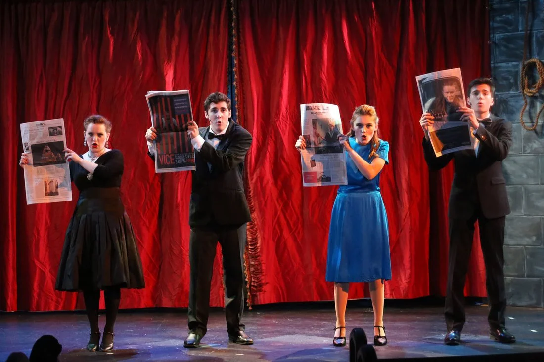 Four actors on stage holding newspapers, standing in front of a red curtain, dressed in formal attire, performing in a theatrical production.