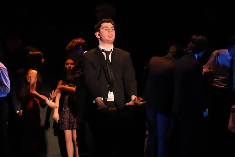 A group of young adults dressed in formal attire, standing and sitting in a dark room with a single light focusing on one young man in the center.