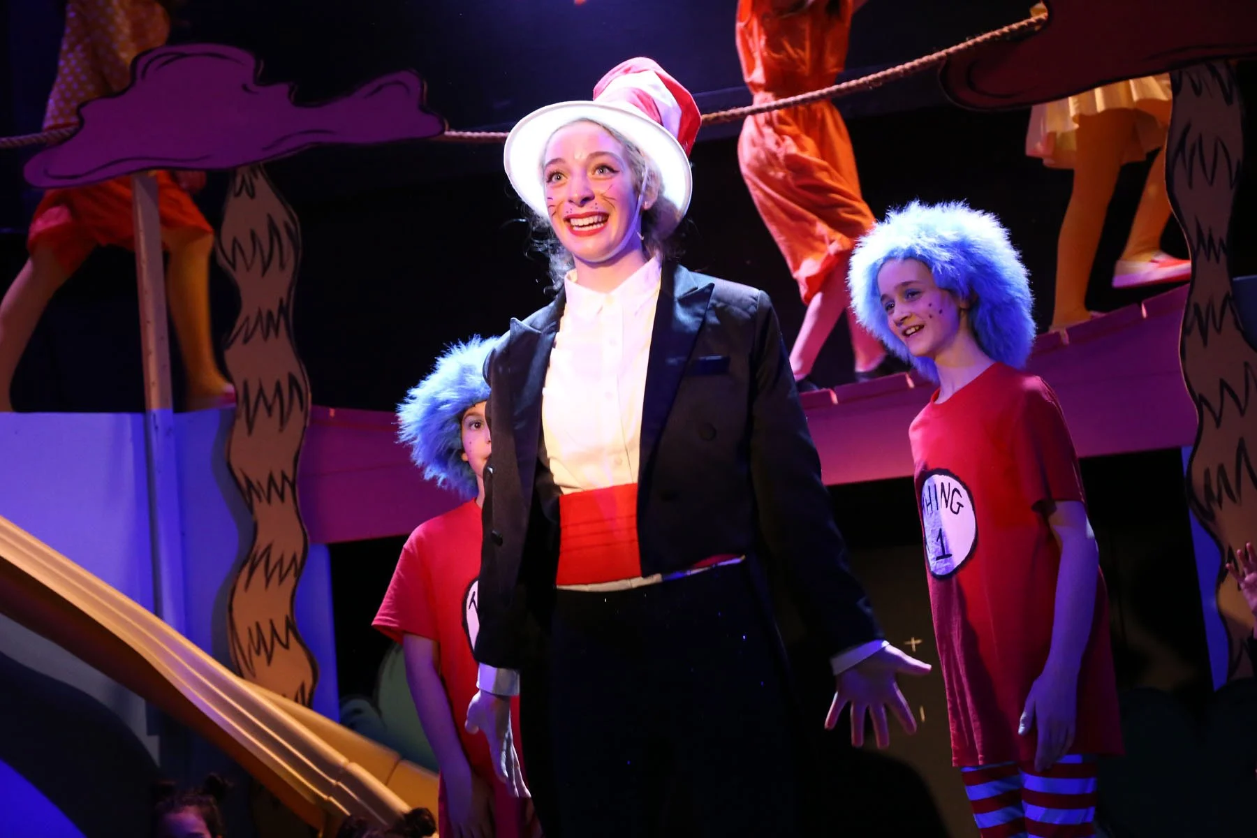 Woman dressed as the Cat in the Hat standing in front of children dressed as Thing 1 and Thing 2 on a stage with colorful Dr. Seuss-themed decorations.