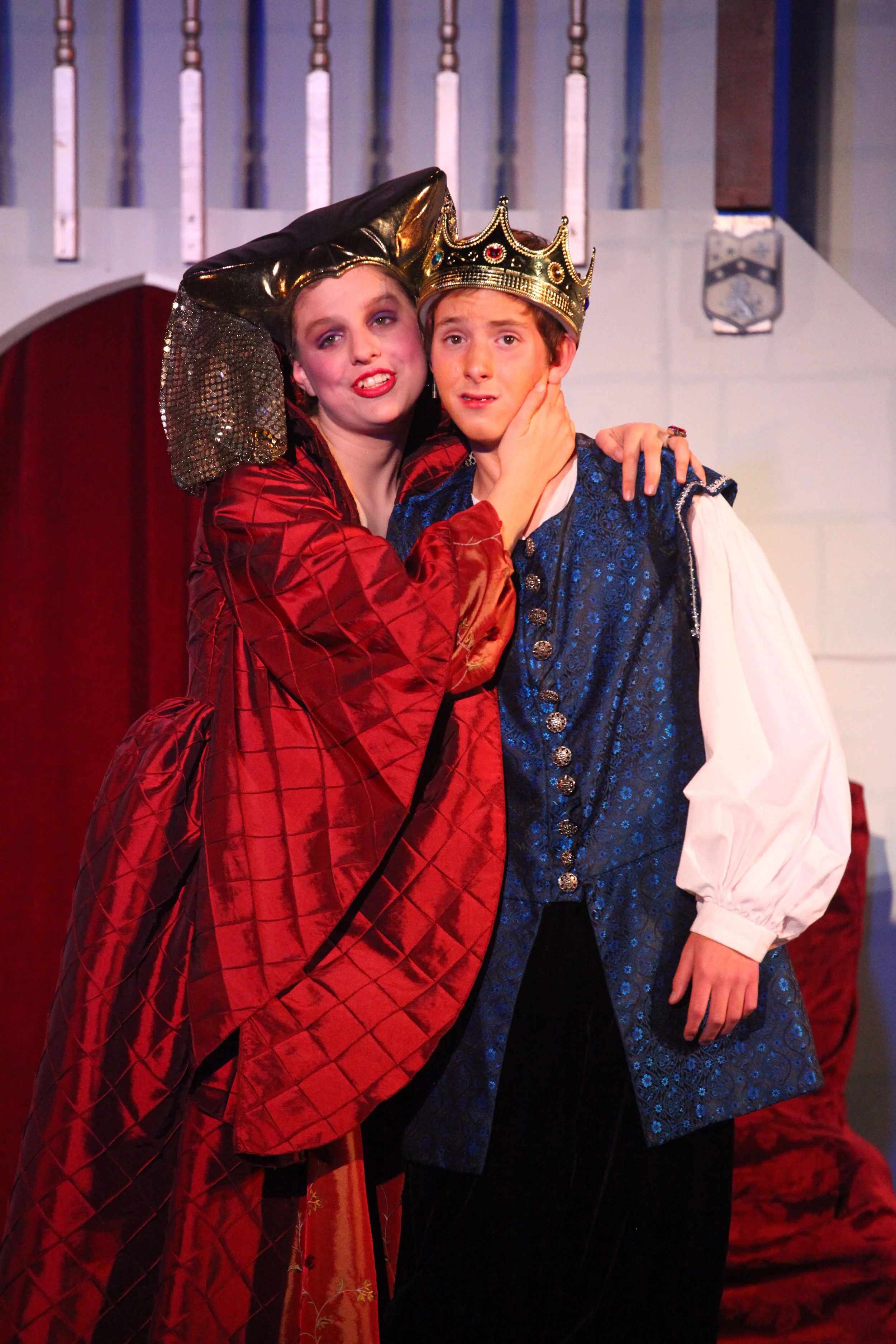 Two actors in royal costumes, one wearing a crown and the other with a jewel-encrusted headpiece, on stage during a theatrical performance.