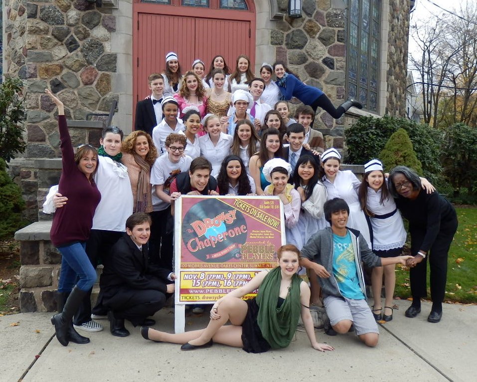 Group of children and teenagers posing in front of a stone building with a red door, for a play called "Drowsy Chaperone".