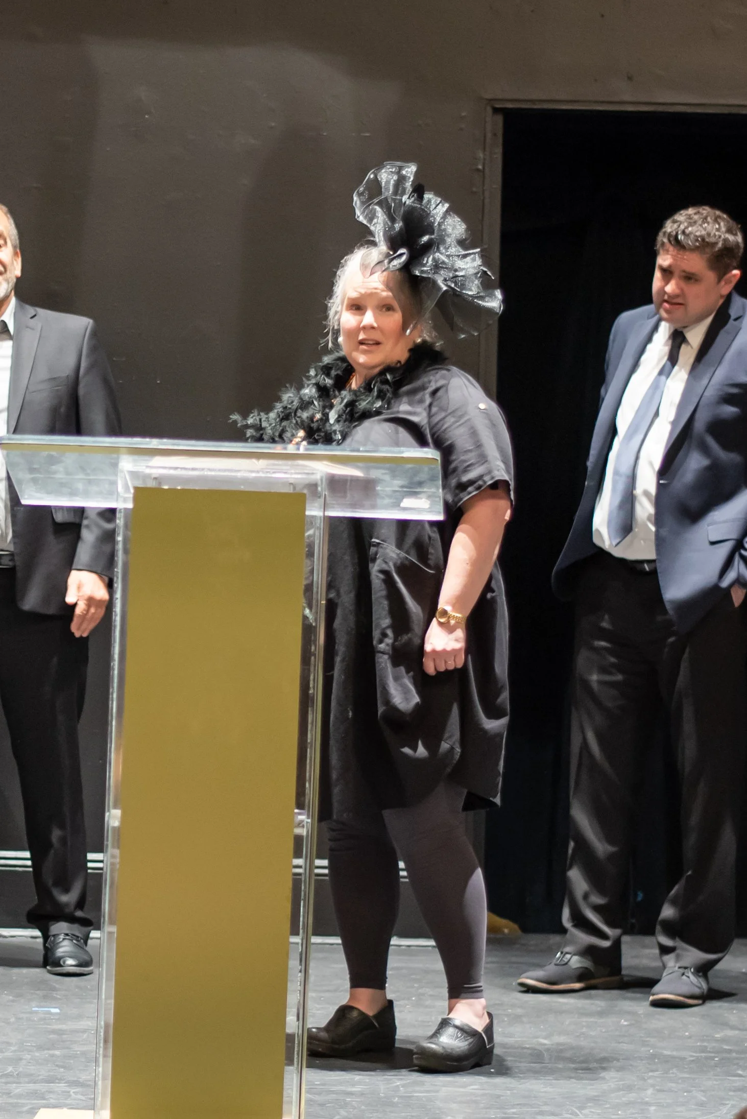 A woman dressed in black with a large black hat and feathered accessory stands behind a transparent podium, speaking into a microphone. Two men in suits stand on either side of her, listening. The setting is Grandma Sylvia's Funeral.