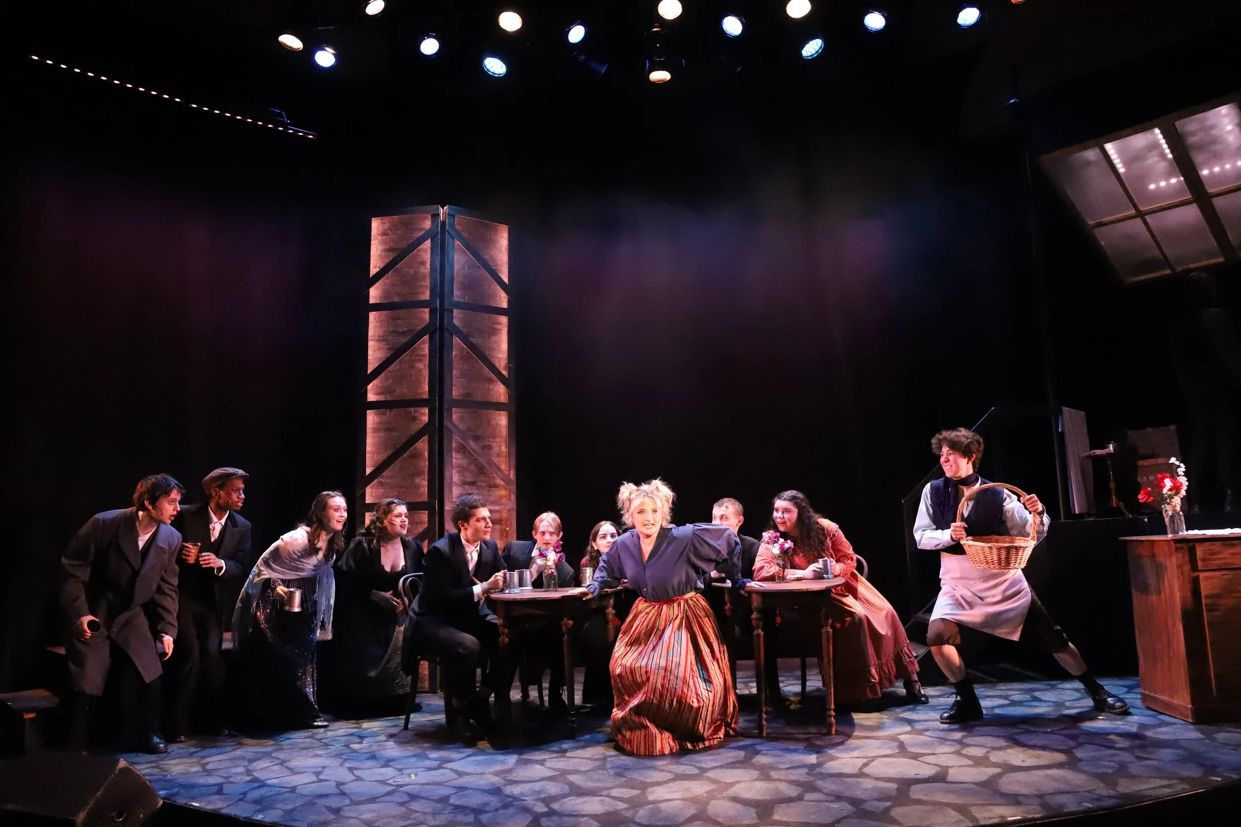 A theatrical stage scene portraying a lively gathering of actors dressed in vintage-style costumes. Several actors sit at and stand around a wooden table, some holding cups, with one woman in a striped skirt and gray blouse at the center appearing an