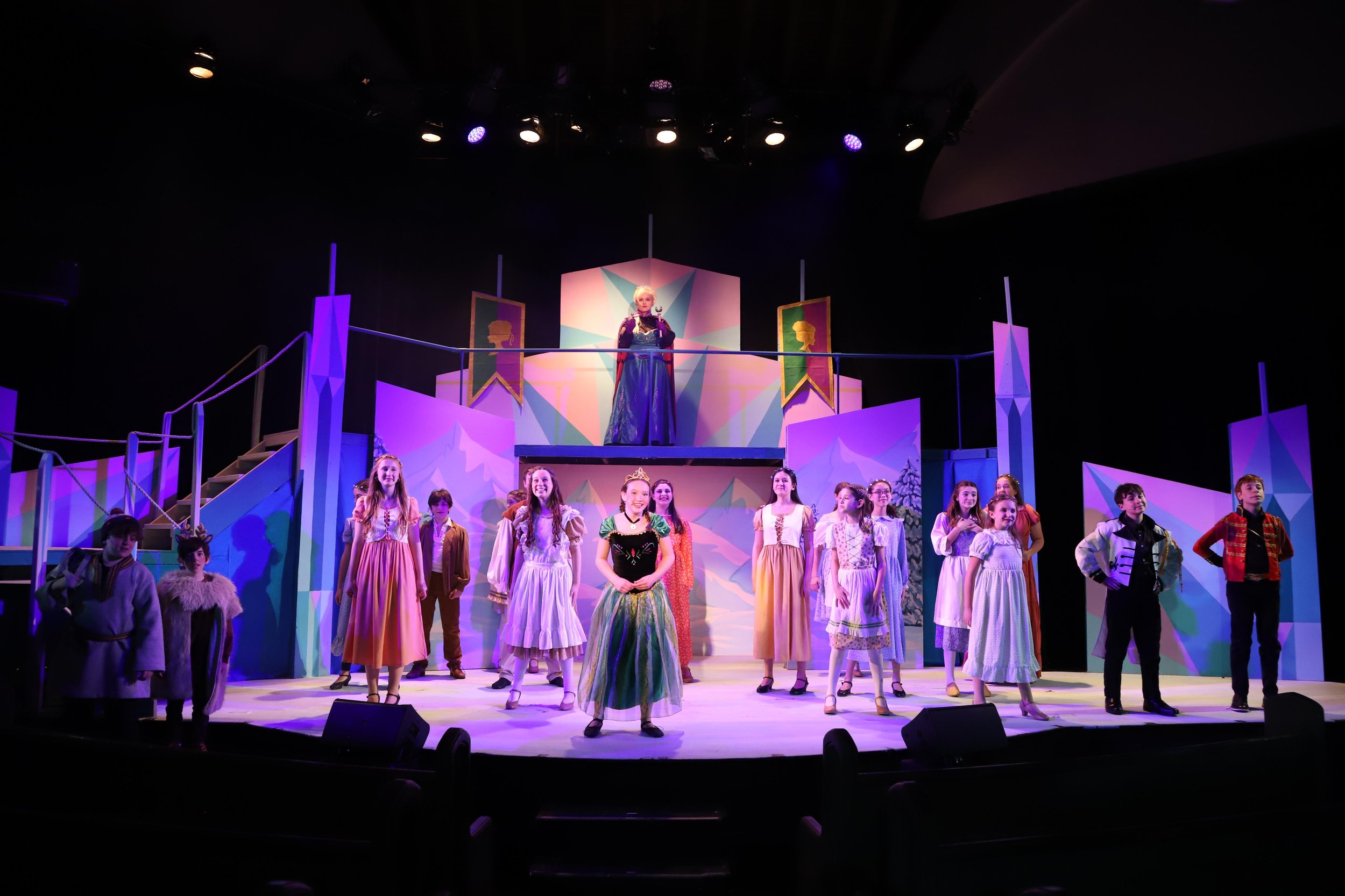 Children dressed as characters from a play standing on stage in a colorful winter scene, with a woman in a royal costume on a raised platform behind them.
