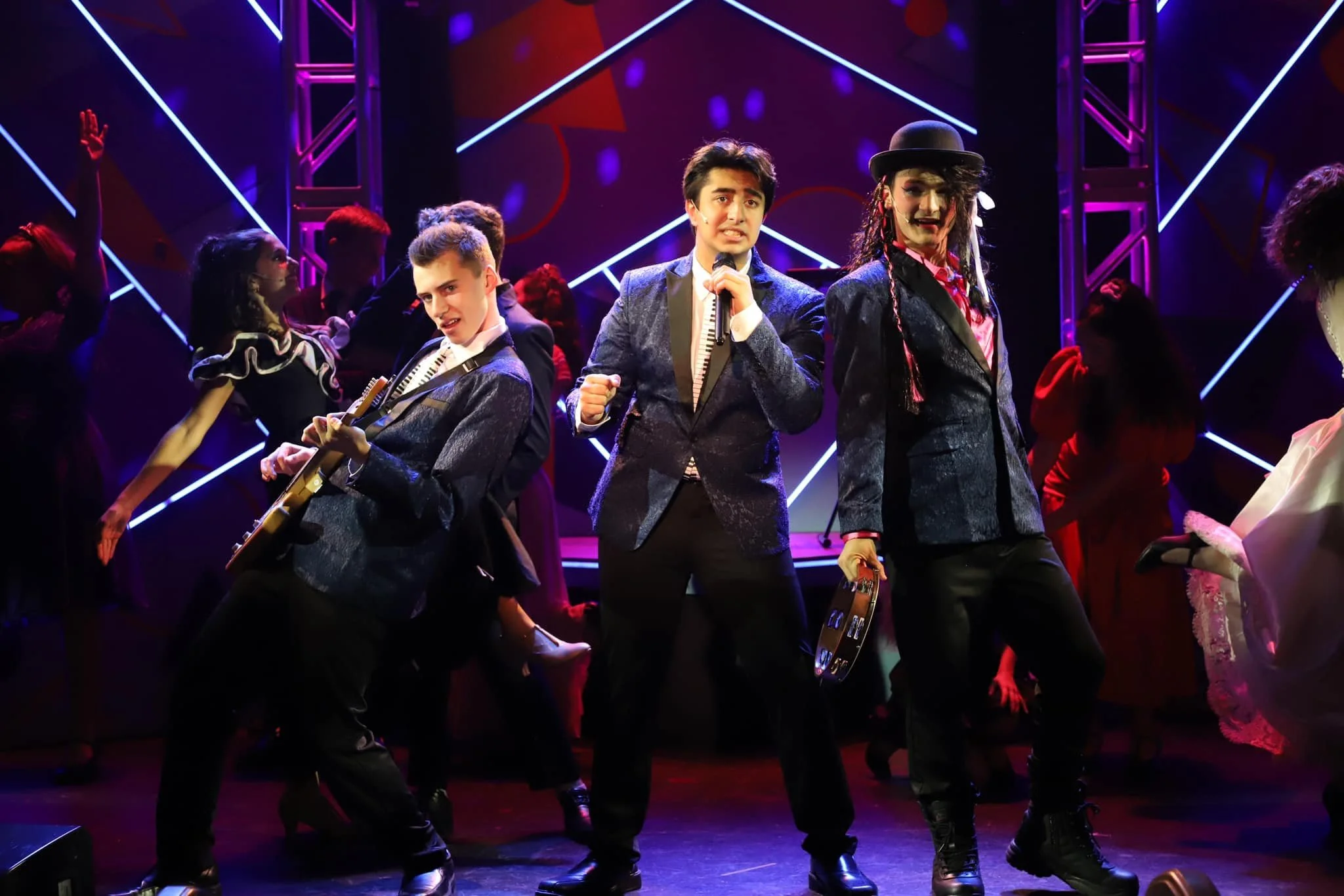 Three young performers on stage in musical costumes, with neon lights and other performers in the background.