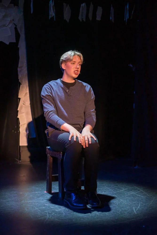 A person sitting on a chair on a stage, performing or speaking, with a black backdrop and some fabric or paper decorations hanging overhead. Performance of Page to Stage cabaret.