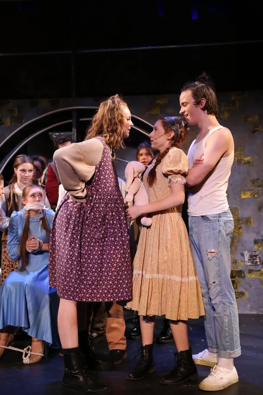 A theatrical scene featuring two women and a young man standing closely and facing each other, with six children watching in the background. The children have various expressions and are dressed in vintage-style clothing. One girl in the background h