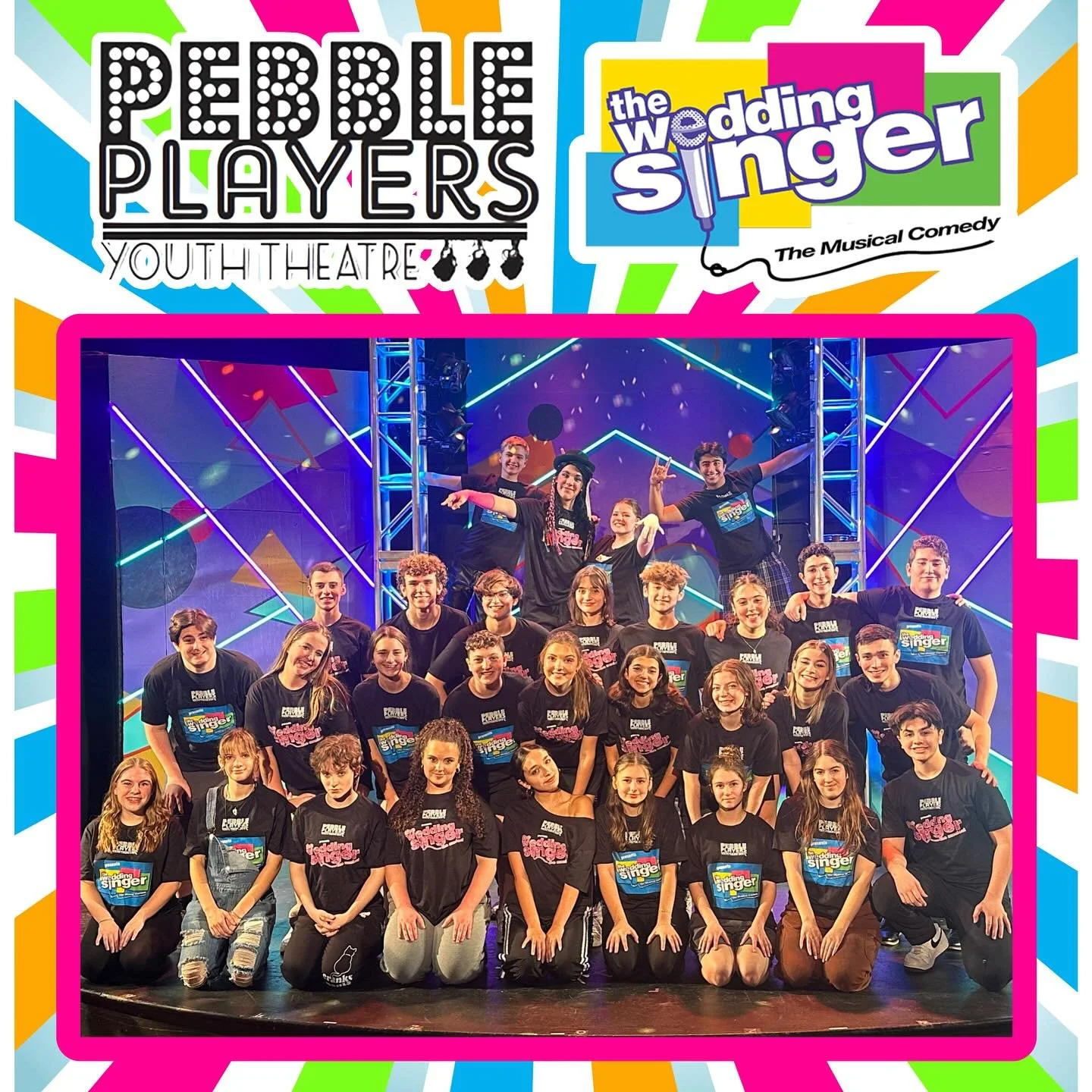 A large group of young performers on stage, posing for a photo after a show, with colorful lighting and geometric background, wearing black t-shirts with the show titles and logos.