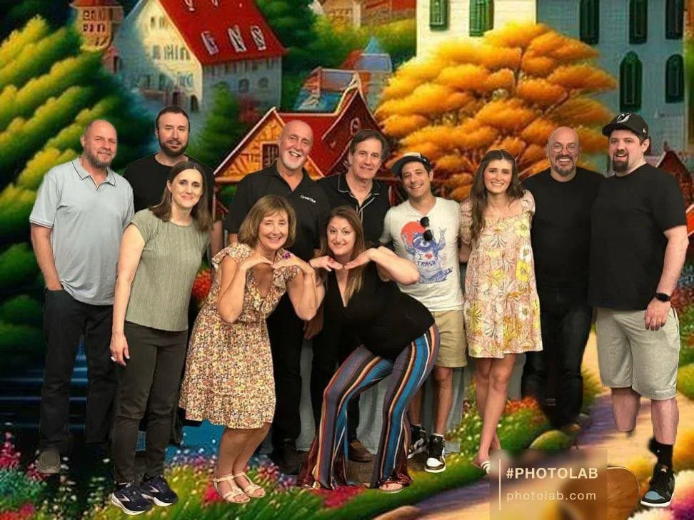 A group of ten people standing together outdoors, smiling, with a colorful, cartoonish background of houses, trees, and a pond.