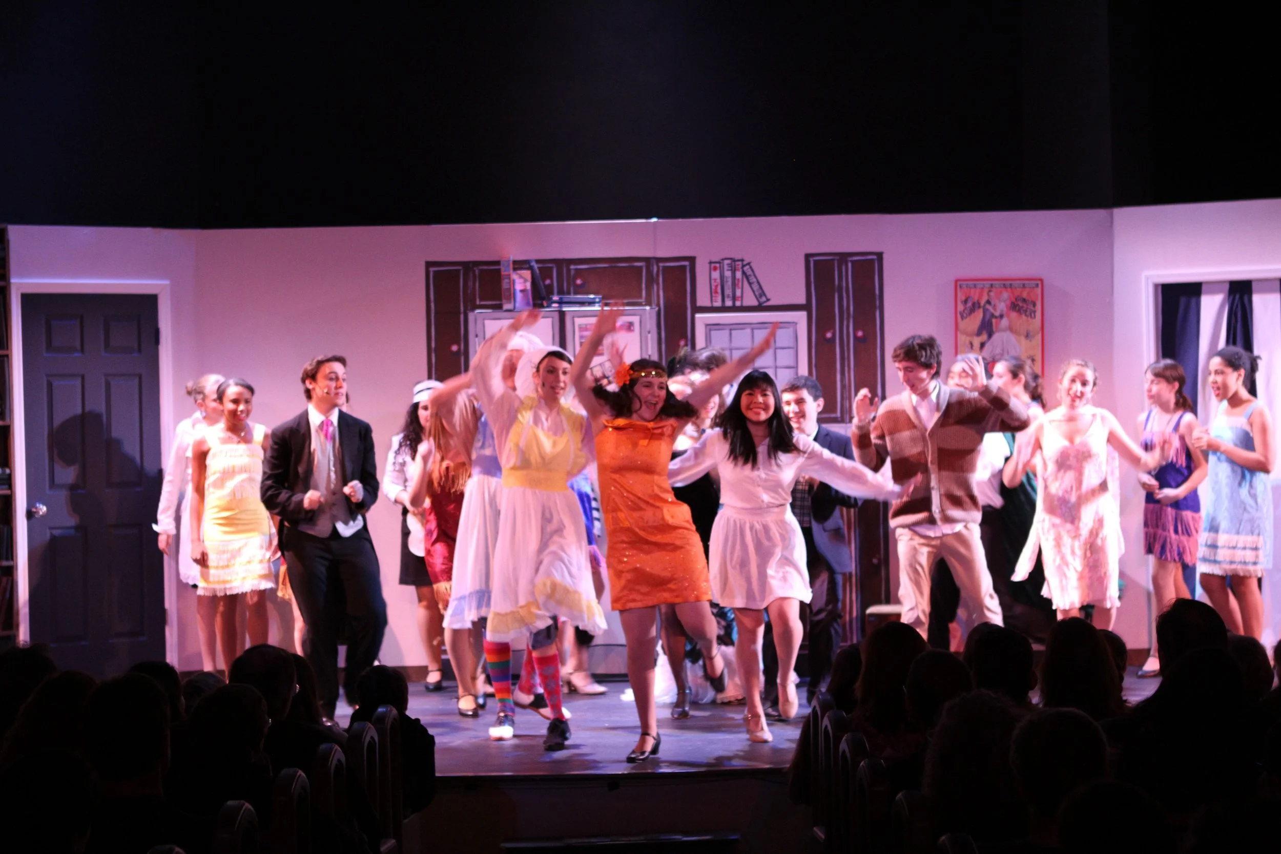 A group of performers on stage during a theatrical production of Drowsy Chaperone, dancing and smiling, with audience members watching in the foreground.