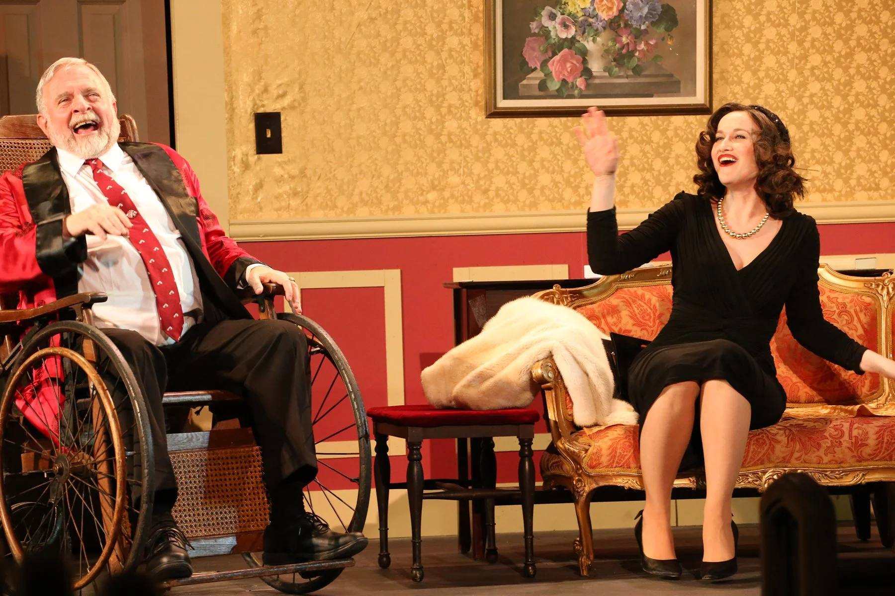 An elderly man in a wheelchair and woman sitting on a vintage sofa, both laughing and engaging in conversation in a warmly decorated room with floral artwork.