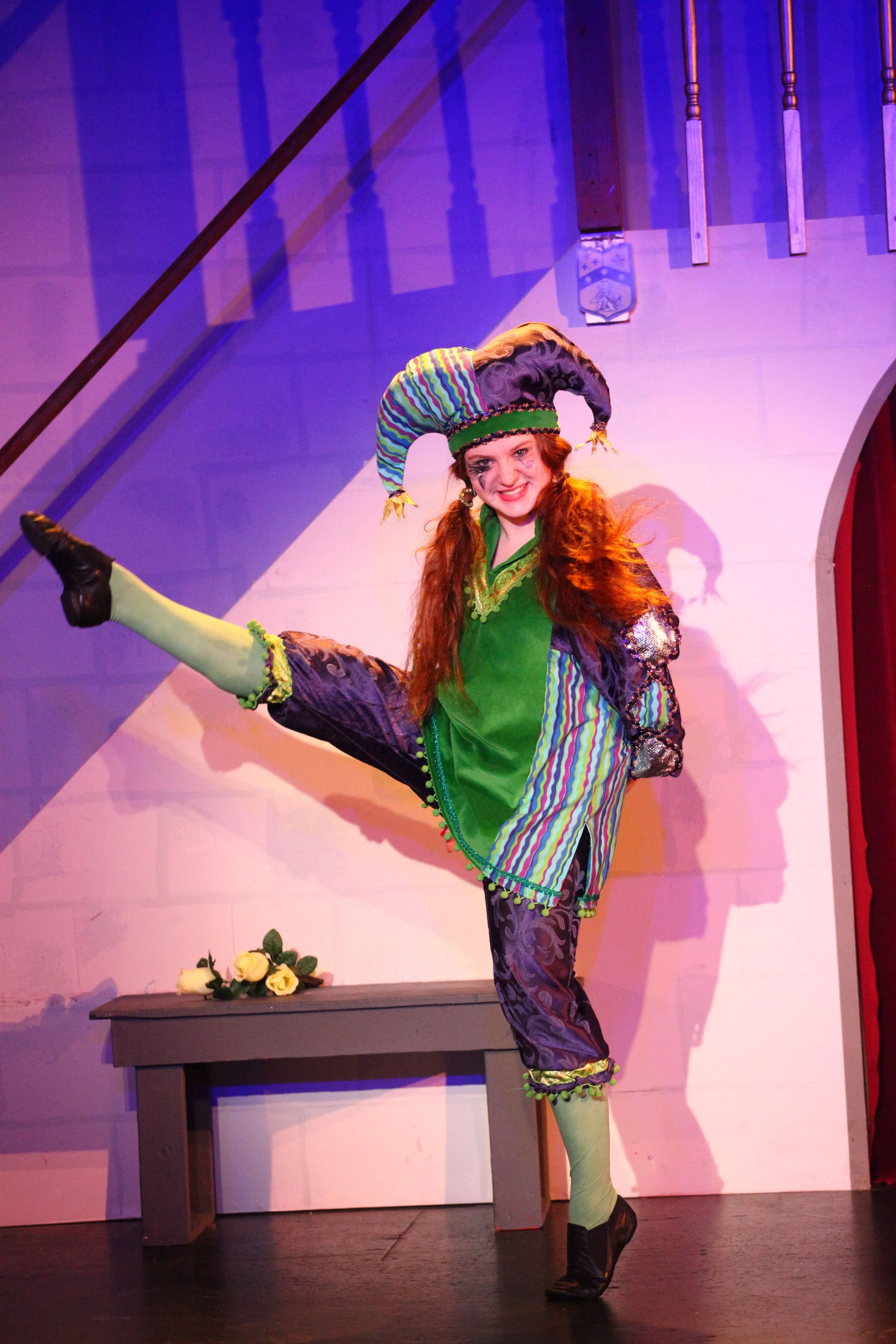 A performer dressed as a jester in colorful costume, balancing on one leg on stage with a raised leg, smiling, with a bench and roses in the background.