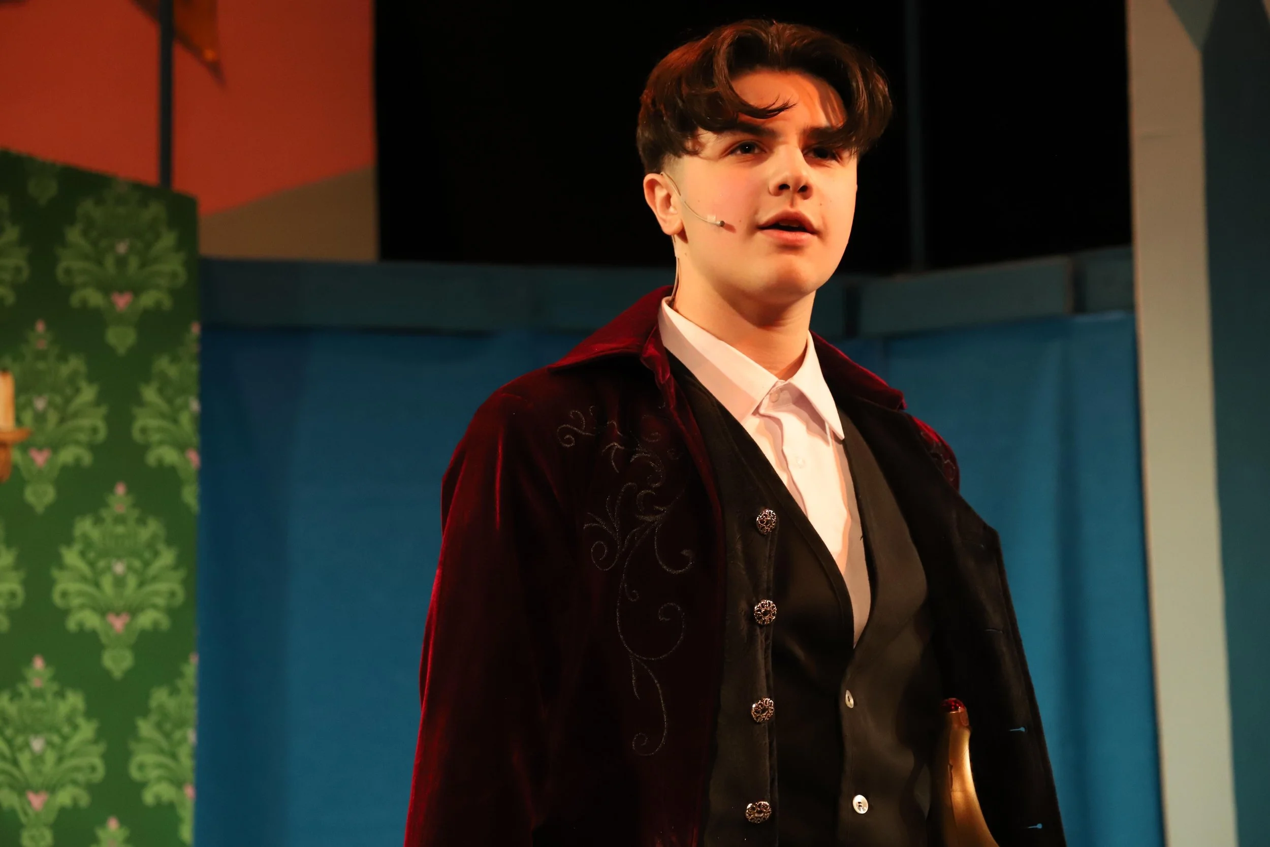 A young man with brown hair wearing a microphone headset, a white collared shirt, a black vest, and a dark red velvet jacket, standing on a colorful stage with blue and green walls.