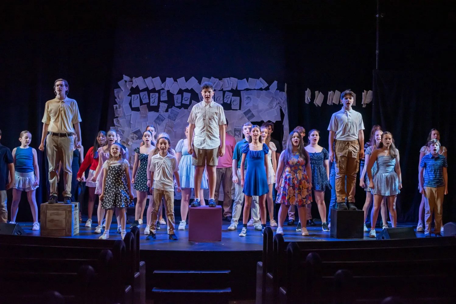 A group of children performing on stage, some standing on wooden blocks, during a theatrical production performance of Page to Stage cabaret.