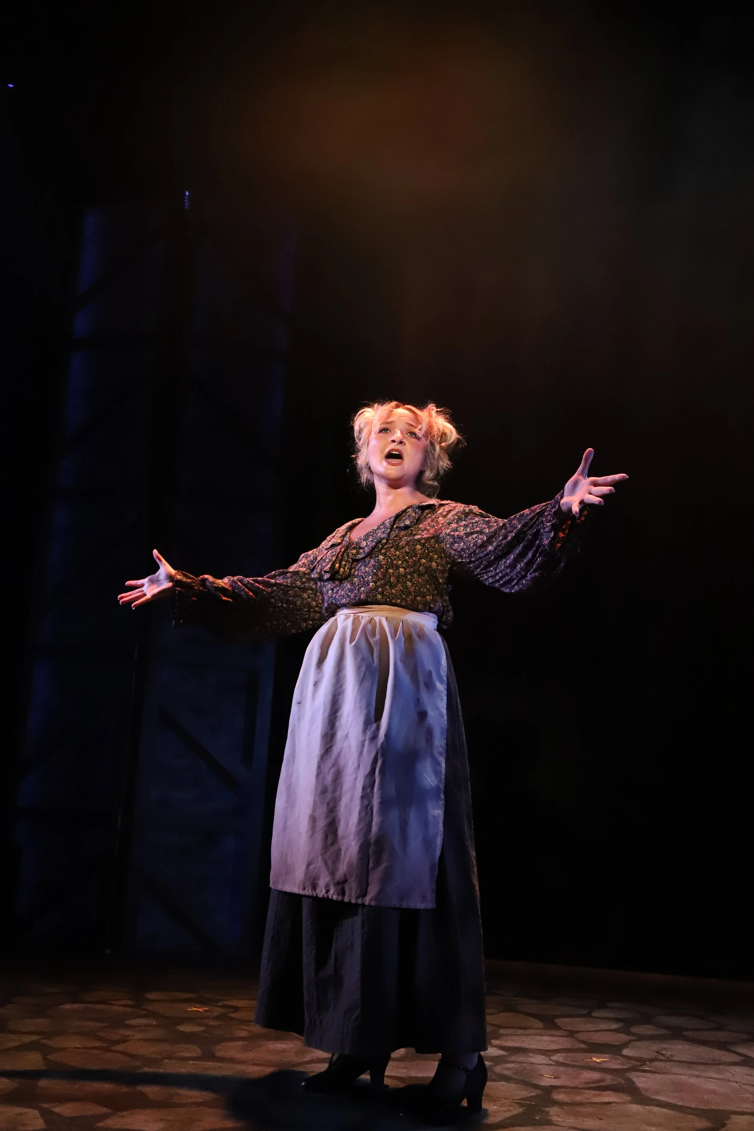 A woman with blonde hair in vintage costume performing on stage with her arms outstretched and mouth open.