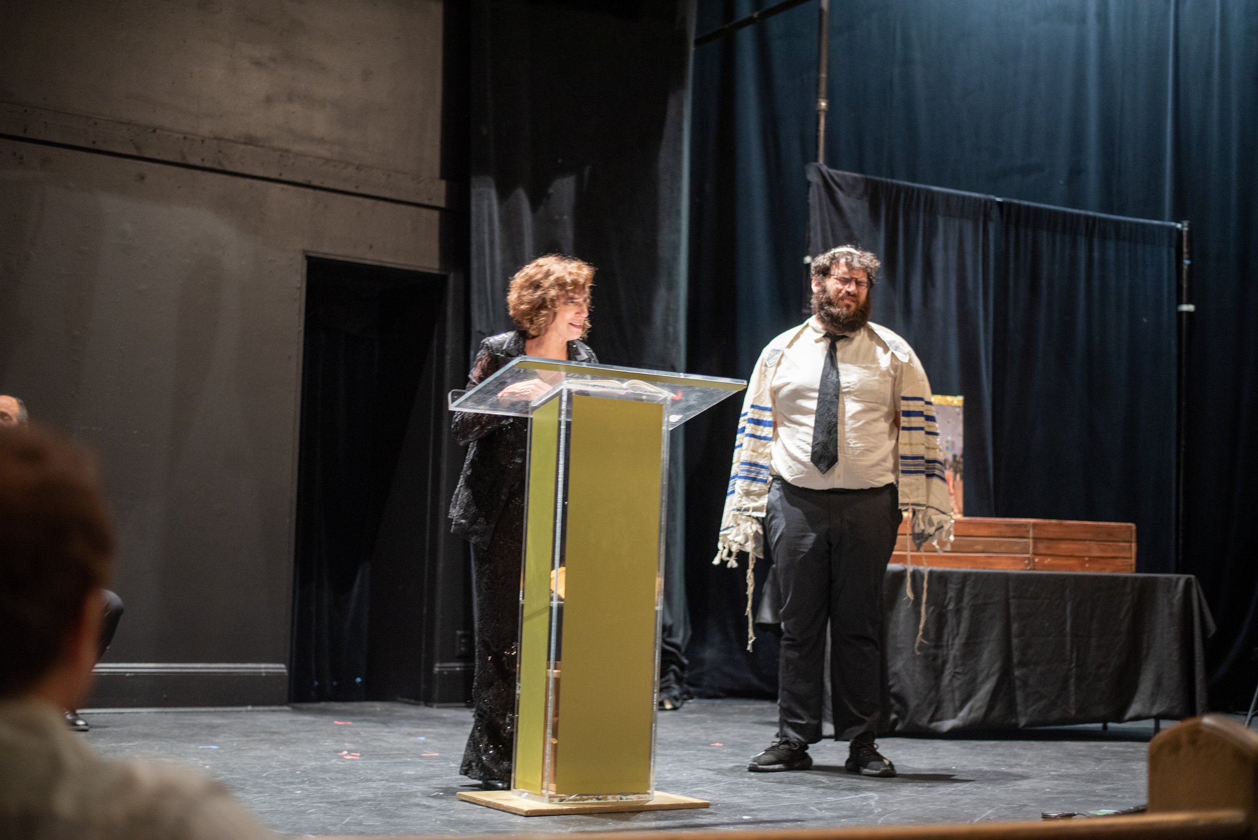 A woman with curly hair standing at a podium speaking and smiling, a man with a beard and glasses standing next to her wearing a white shirt with a shawl, black pants, and a black tie, and a table with a coffin behind them.