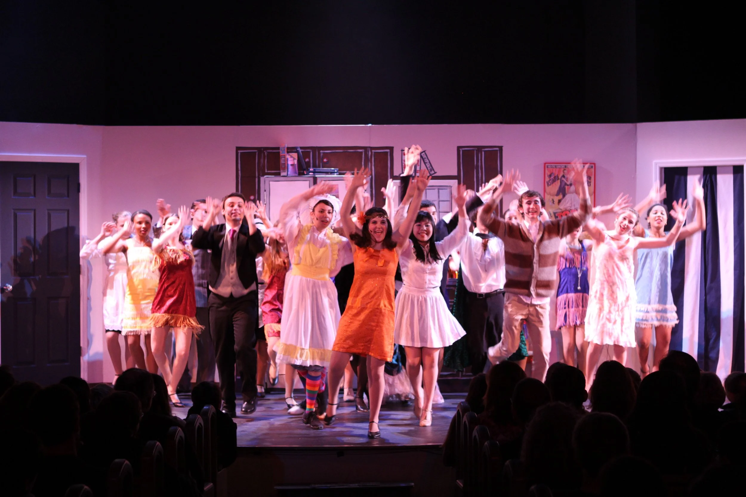 The cast of a musical or play performing on stage of Drowsy Chaperone with a set resembling a room, including props and posters, while audience members watch.