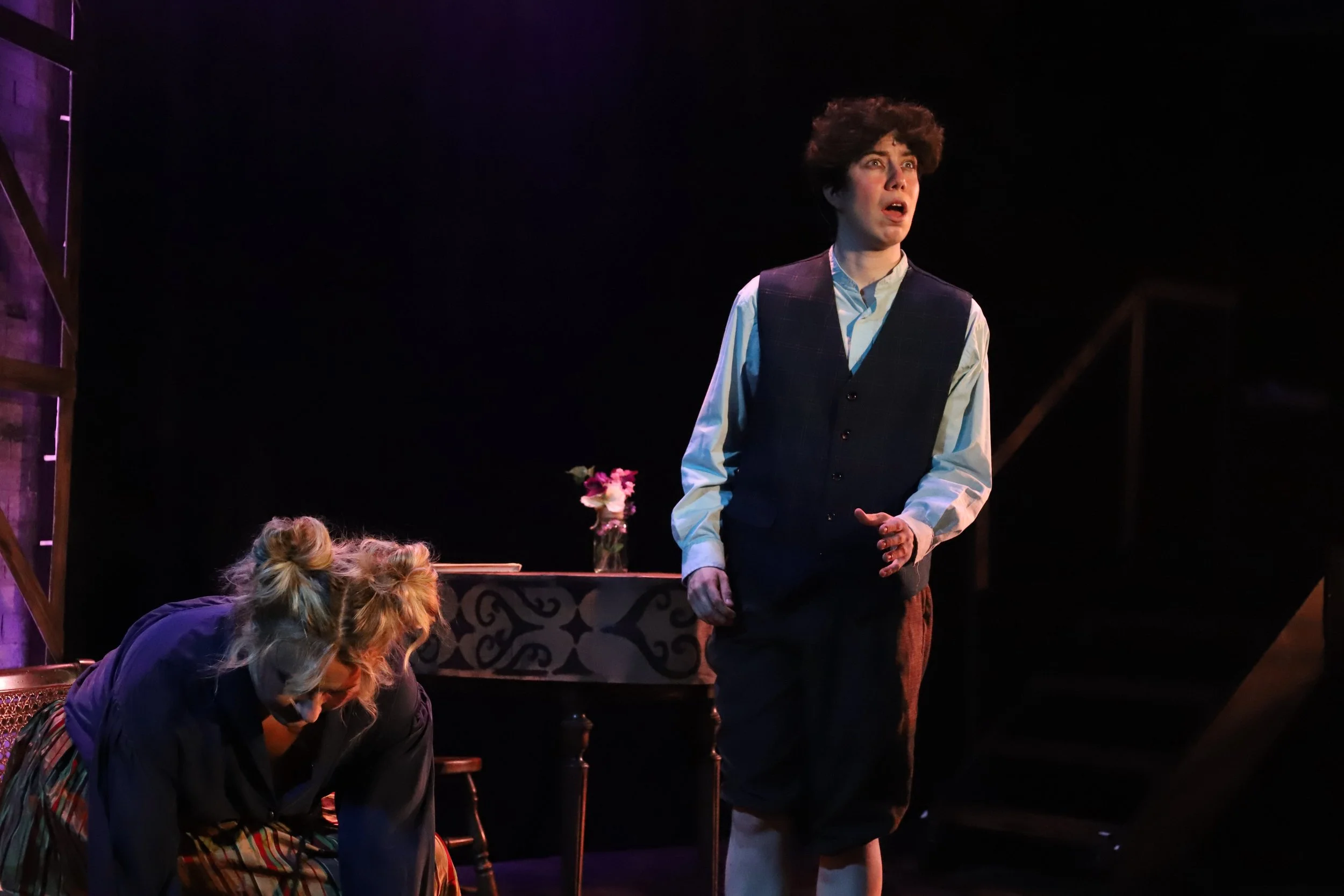 Two actors performing on stage, one standing with a surprised expression and the other kneeling or bent over with their head down. A table with a vase of flowers is in the background.