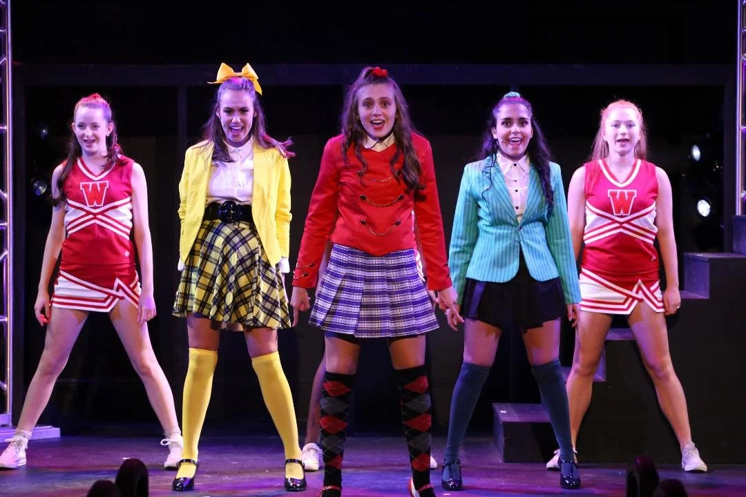 Group of young girls performing on stage in colorful school-themed costumes, with three in cheerleader outfits and two in school uniform styles.