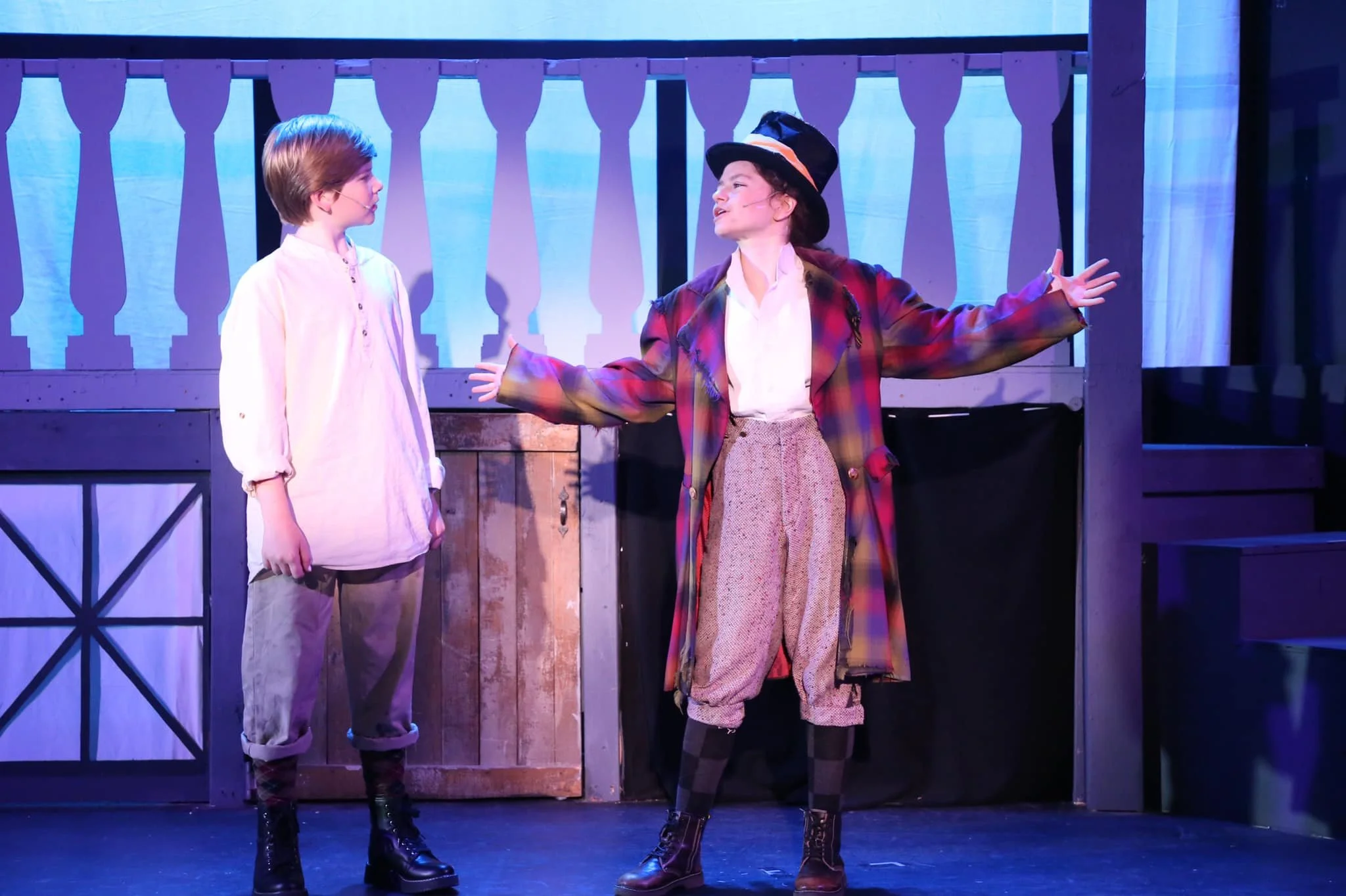 Two actors on stage performing in a play, one young boy dressed in period costume and an actress dressed as a witch, with a wooden barn backdrop.