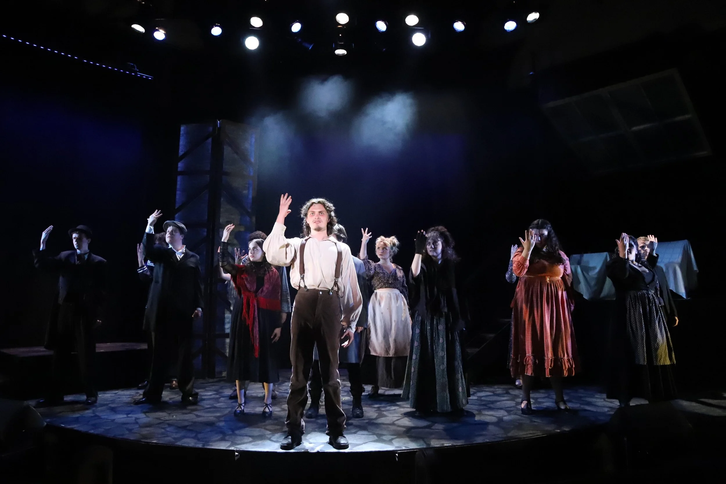 A theatrical performance on stage with a man at the front raising his hand, surrounded by cast members in period costumes, under stage lighting.