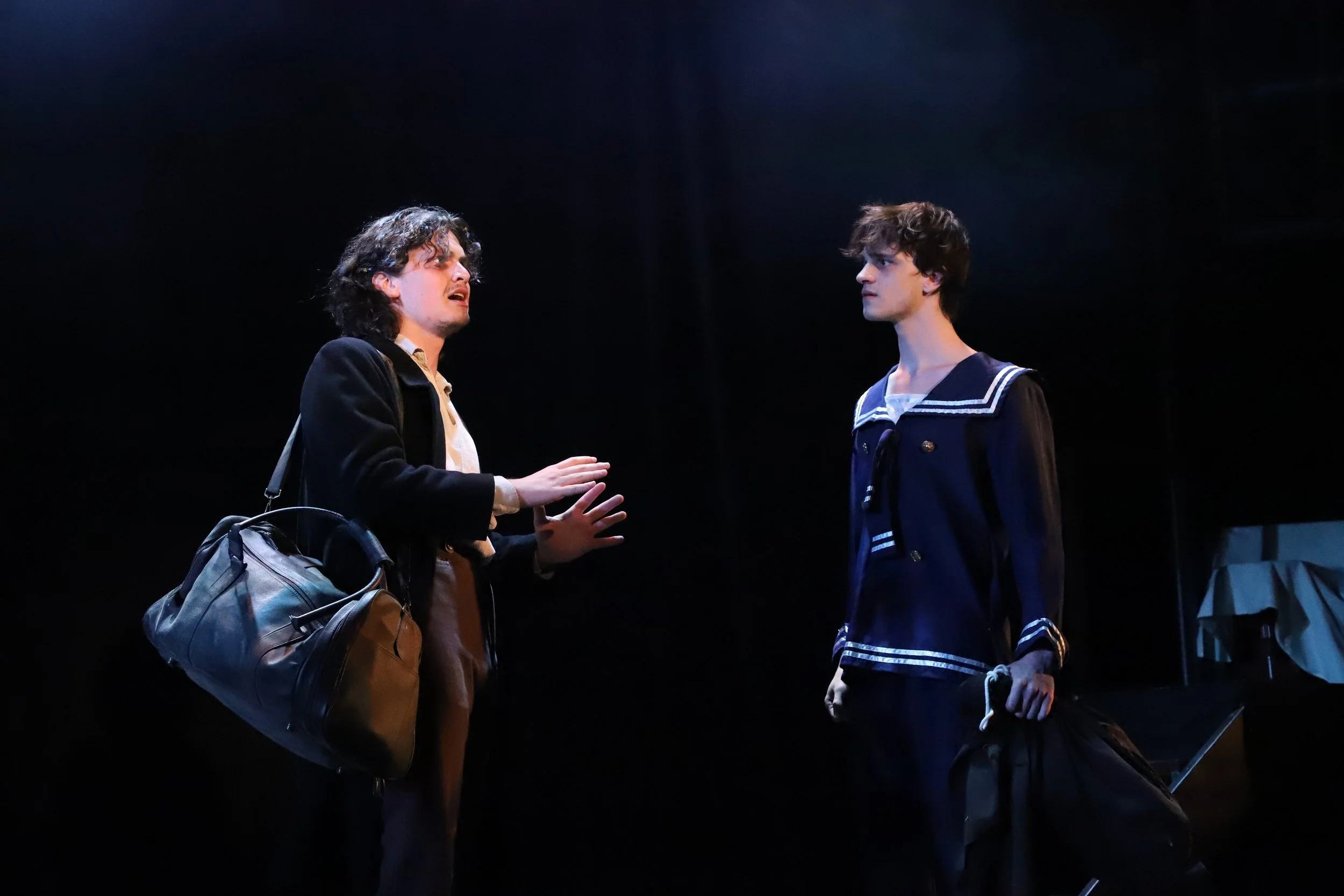 Two young men engaged in a dramatic conversation on stage, one with curly hair and a bag, the other in a navy sailor suit, in a theatrical performance.
