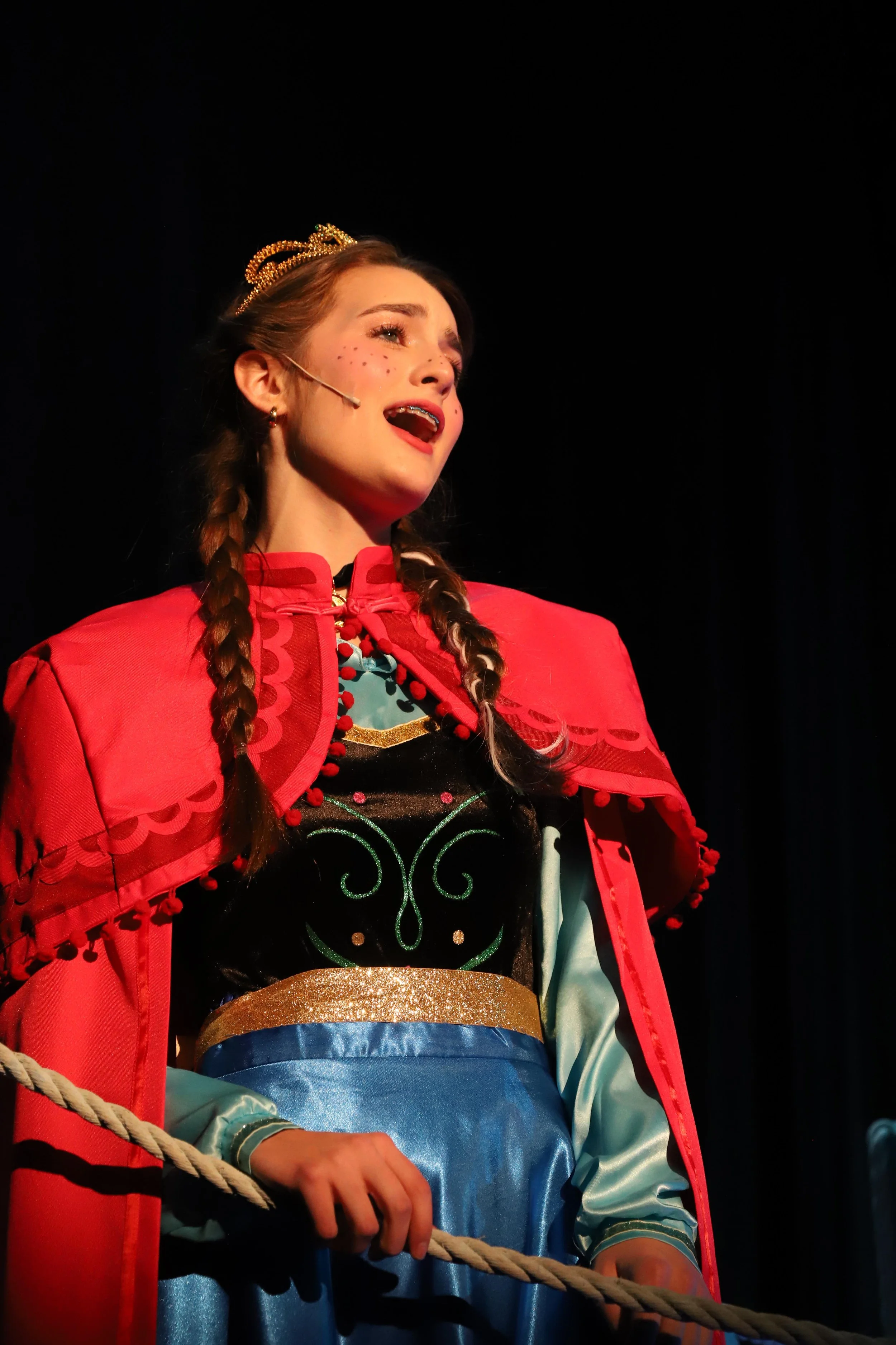 A young woman performing on stage dressed as Anna in Frozen, wearing a red cape, princess gown, and tiara, with an emotional expression.