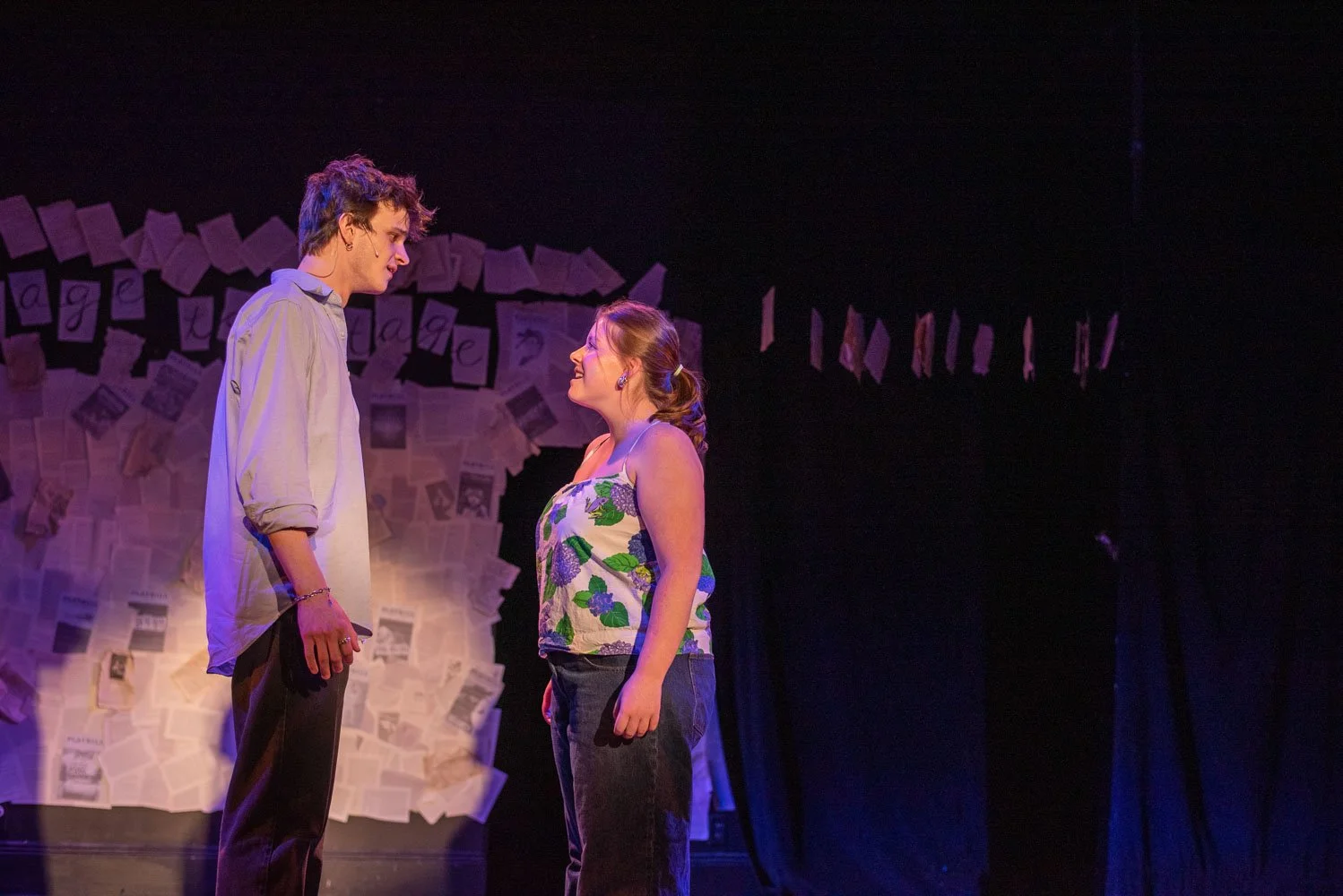 Two young actors, a male and a female, stand face to face on stage, engaged in a scene. The male has curly hair and is wearing a light-colored shirt, while the female has long hair tied back and is wearing a floral sleeveless top. The background feat