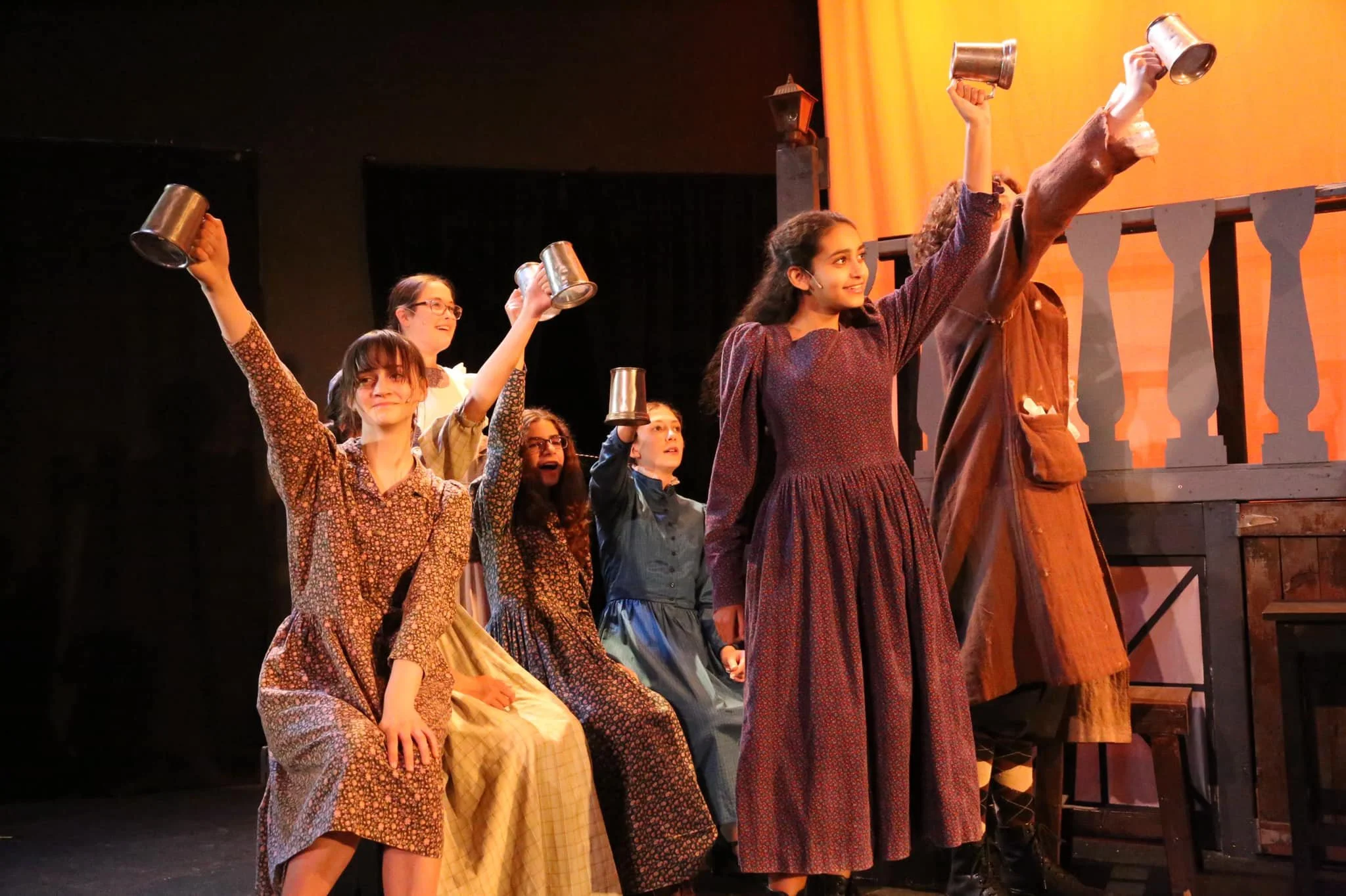 Group of young girls and women on stage during a play, raising cups in celebration, dressed in period costumes, with orange backdrop and dark stage surroundings.