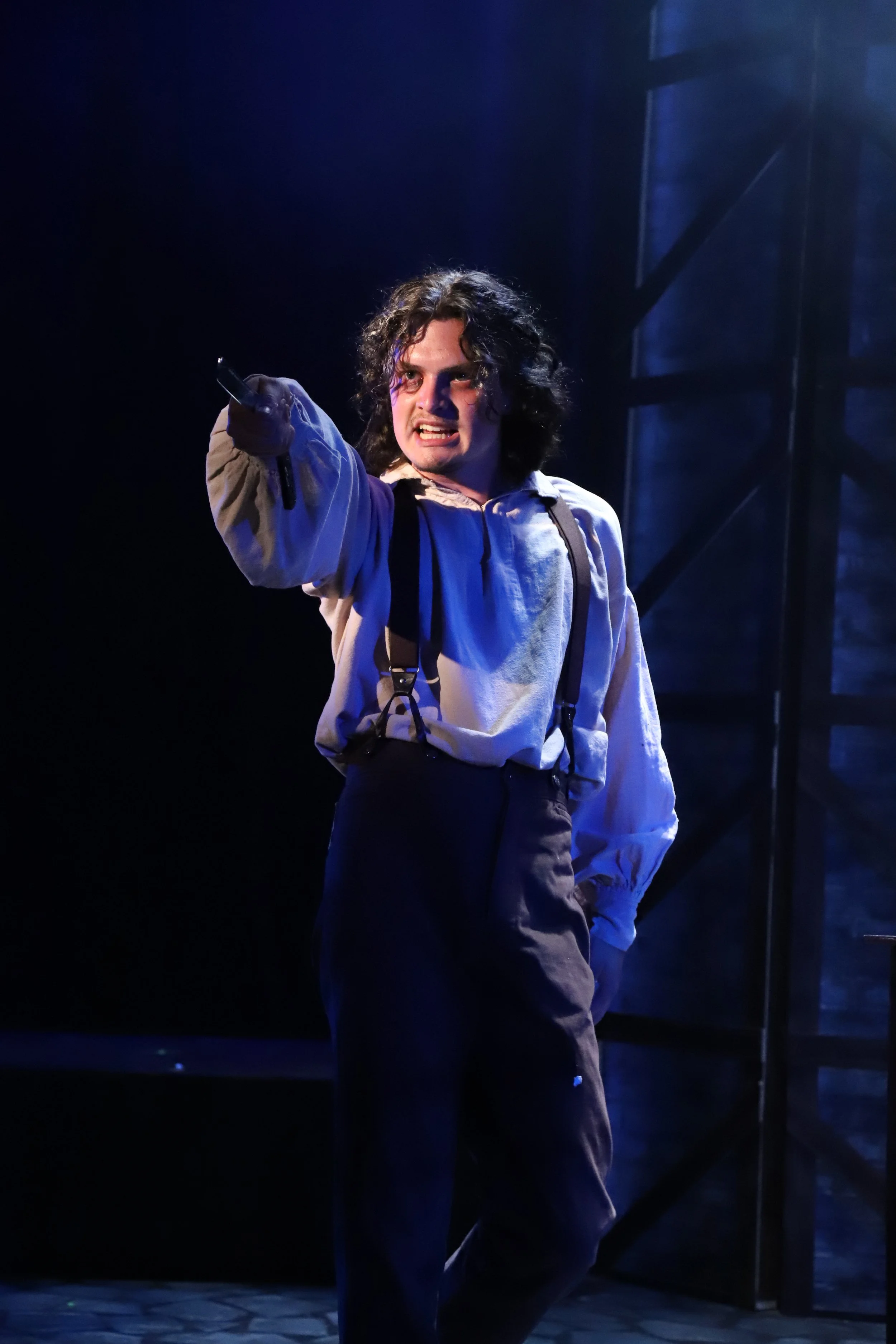 A performer with curly hair and a dramatic expression, dressed in a vintage style shirt and suspenders, stage lighting in blue and purple, theatrical background.