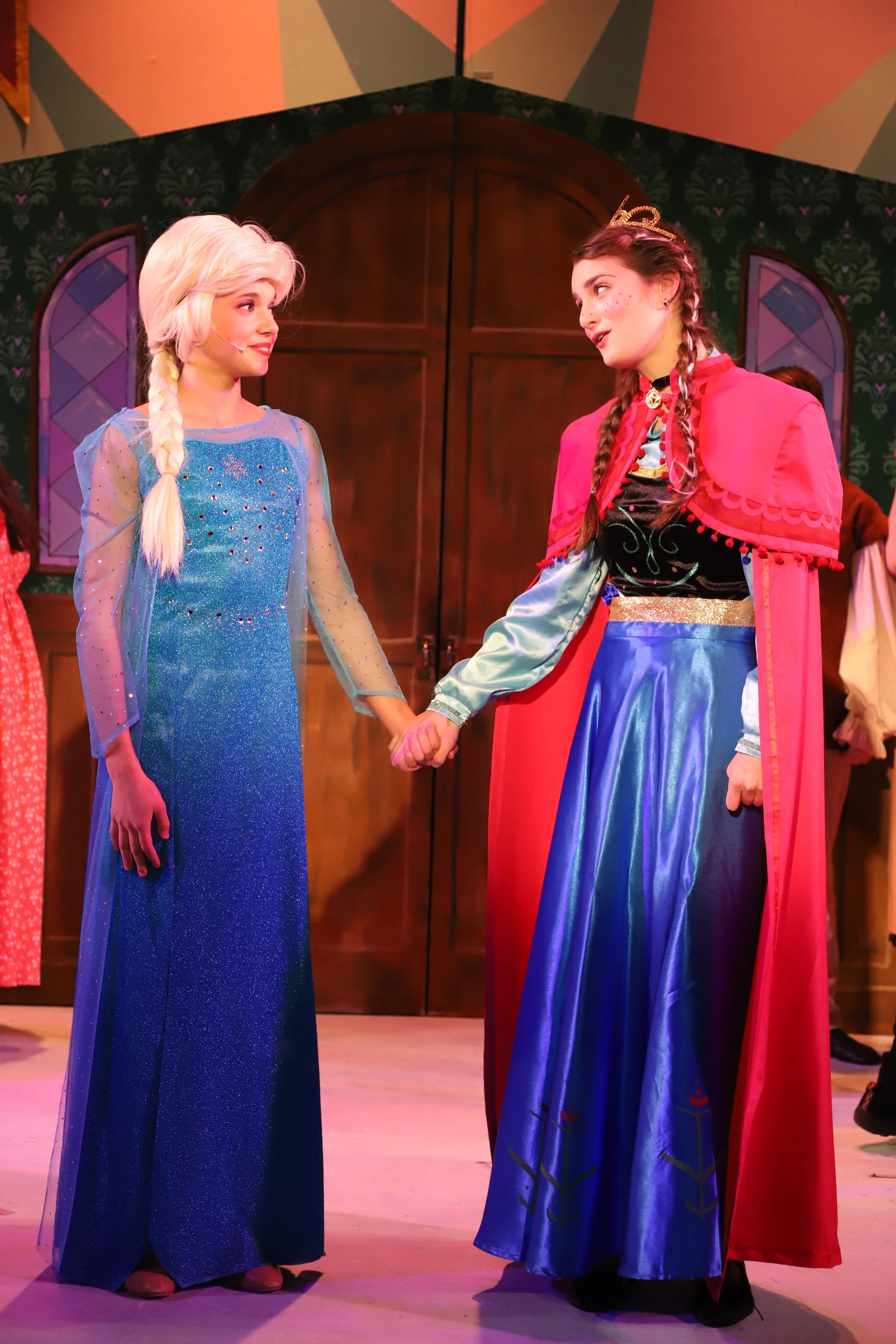 Two young women are holding hands in a scene from Disney's Frozen Jr. Elsa, on the left, has long platinum blonde hair in a braid, a sparkly blue gown, and a sheer cape. Anna, on the right, has brown hair in braids, a red cape, and a blue skirt. 
