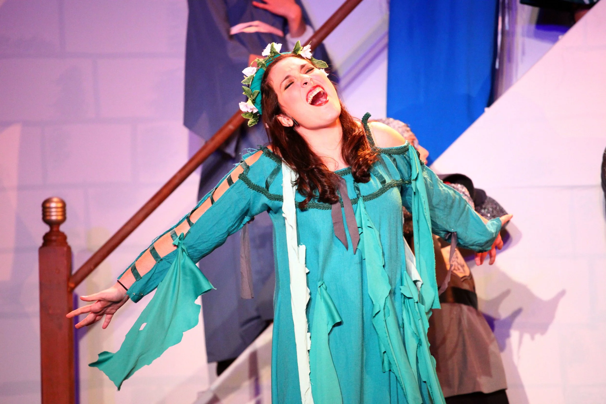 A woman in a blue costume with green leaves and white flowers on her head, singing with her eyes closed and arms outstretched, on stage.