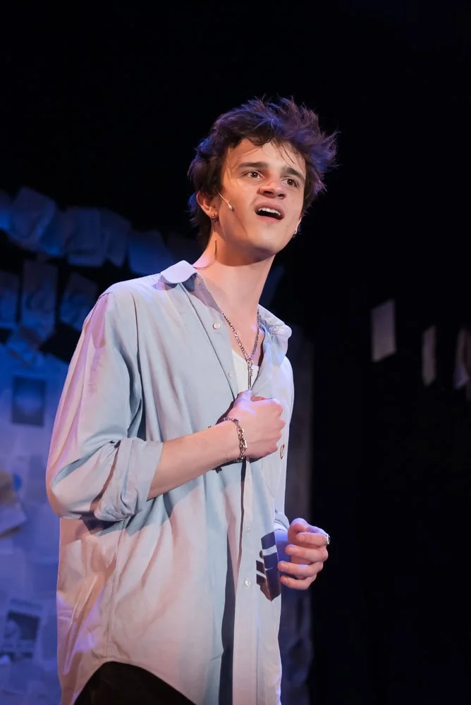 A young man performing on stage with a distressed expression, wearing a light-colored open shirt and a necklace. Performance of Page to Stage cabaret.