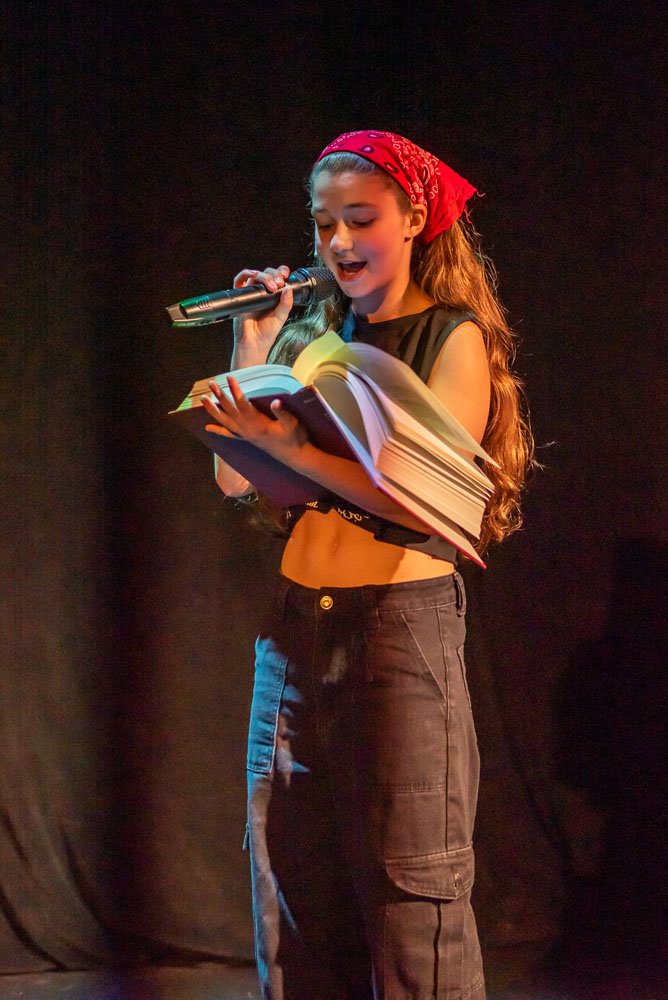 A young girl with long hair, wearing a red bandana, black sleeveless top, and black pants, is reading from a book while speaking into a microphone on a dark stage. Performance of Page to Stage cabaret.
