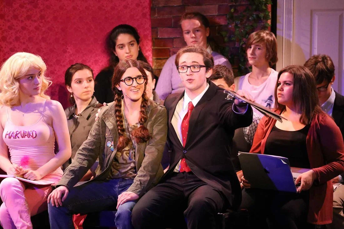 A group of young people sitting in a room, with a boy in glasses and a suit gesturing as if speaking, surrounded by others who appear to be listening.