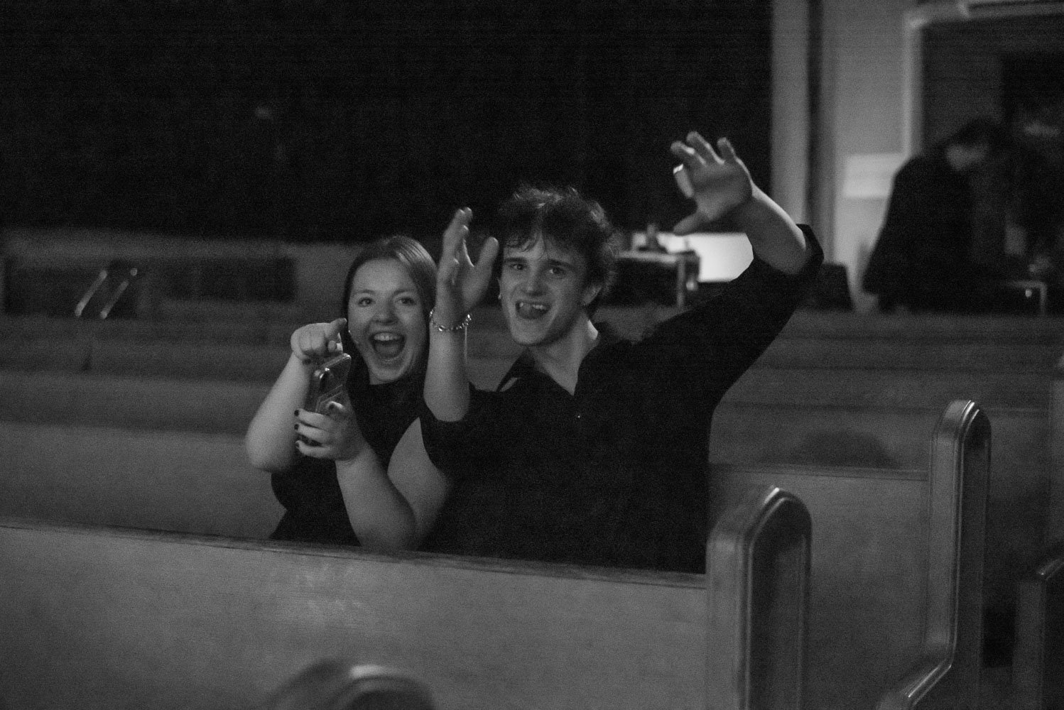 Black and white photo of a young woman and man smiling and waving, sitting in wooden pews in a church at night. Performance of Page to Stage cabaret.