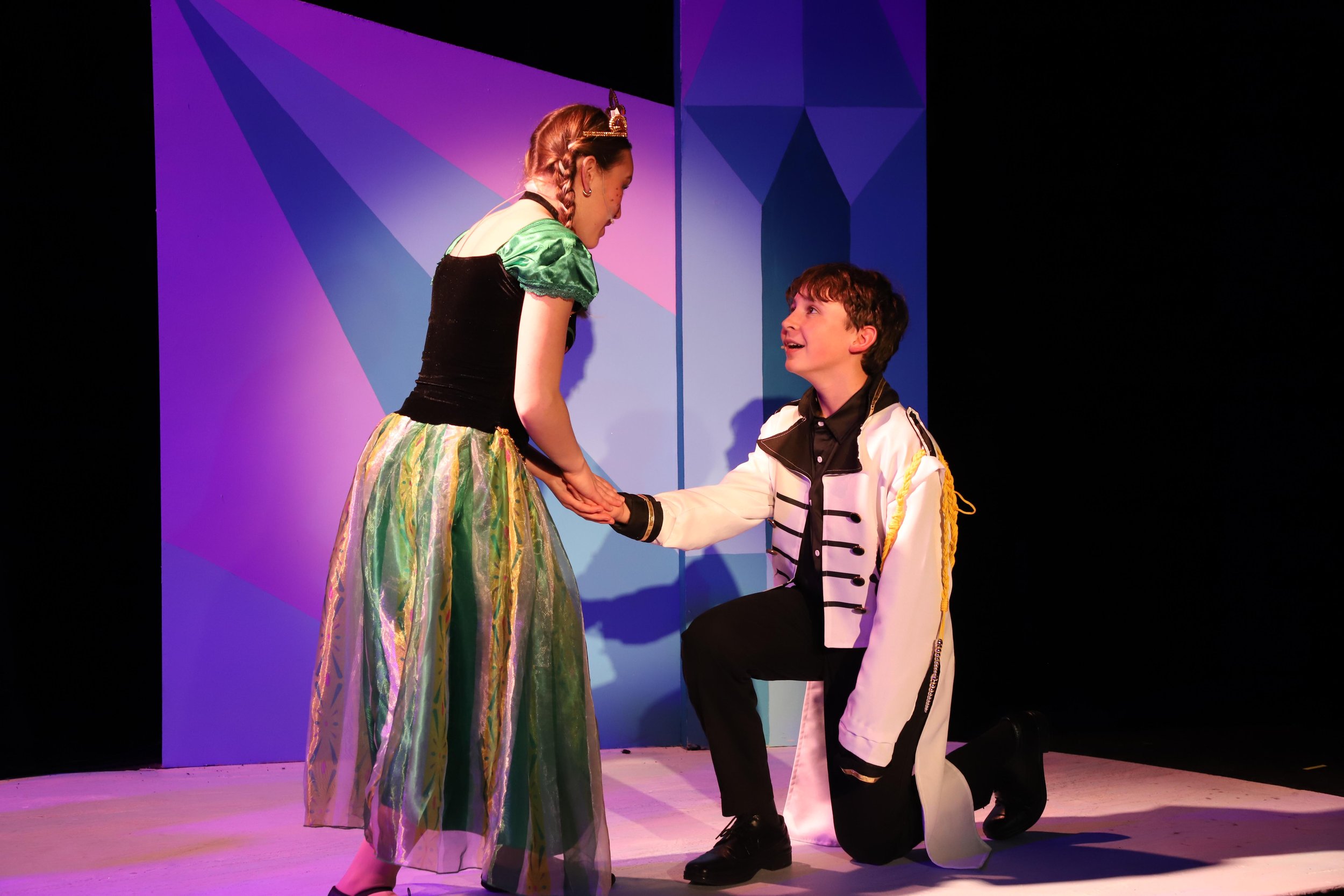 Two children dressed in costumes performing on stage; the girl is wearing a green and black dress with a tiara, and the boy is dressed as a prince in a white coat with gold accents, kneeling with his hand held by the girl.