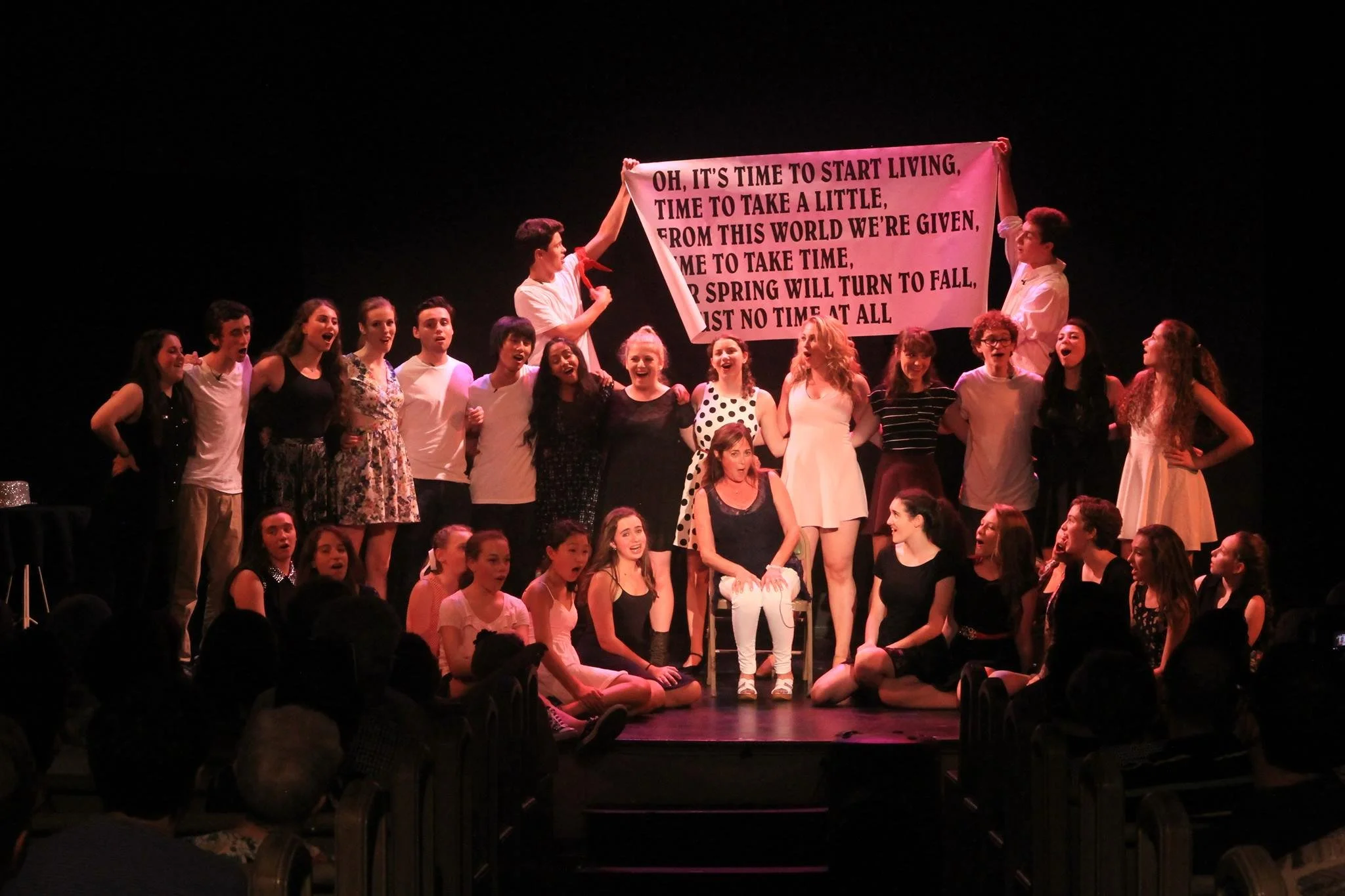 Group of young people on stage, with two holding a large pink sign with lyrics, performing in a theater setting. Scene from 100 Years of Broadway.  
