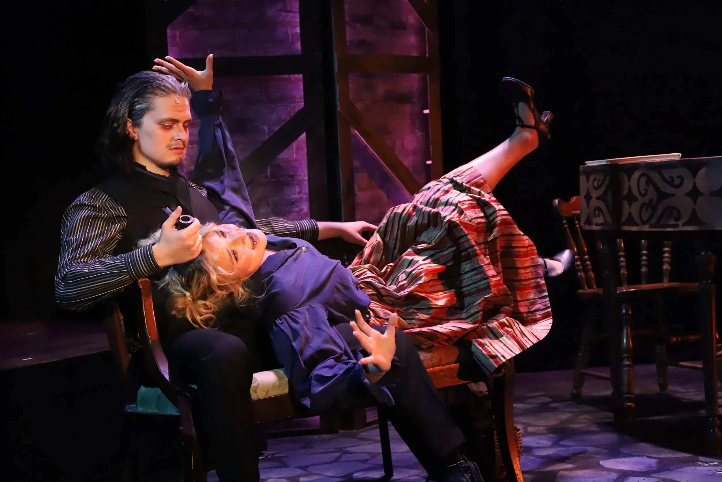 A theatrical scene with two actors on stage. The male actor is seated, with long dark hair, a black and striped costume, holding a small object near the face of the female actress. The female actress is lying across the male's lap, looking distressed