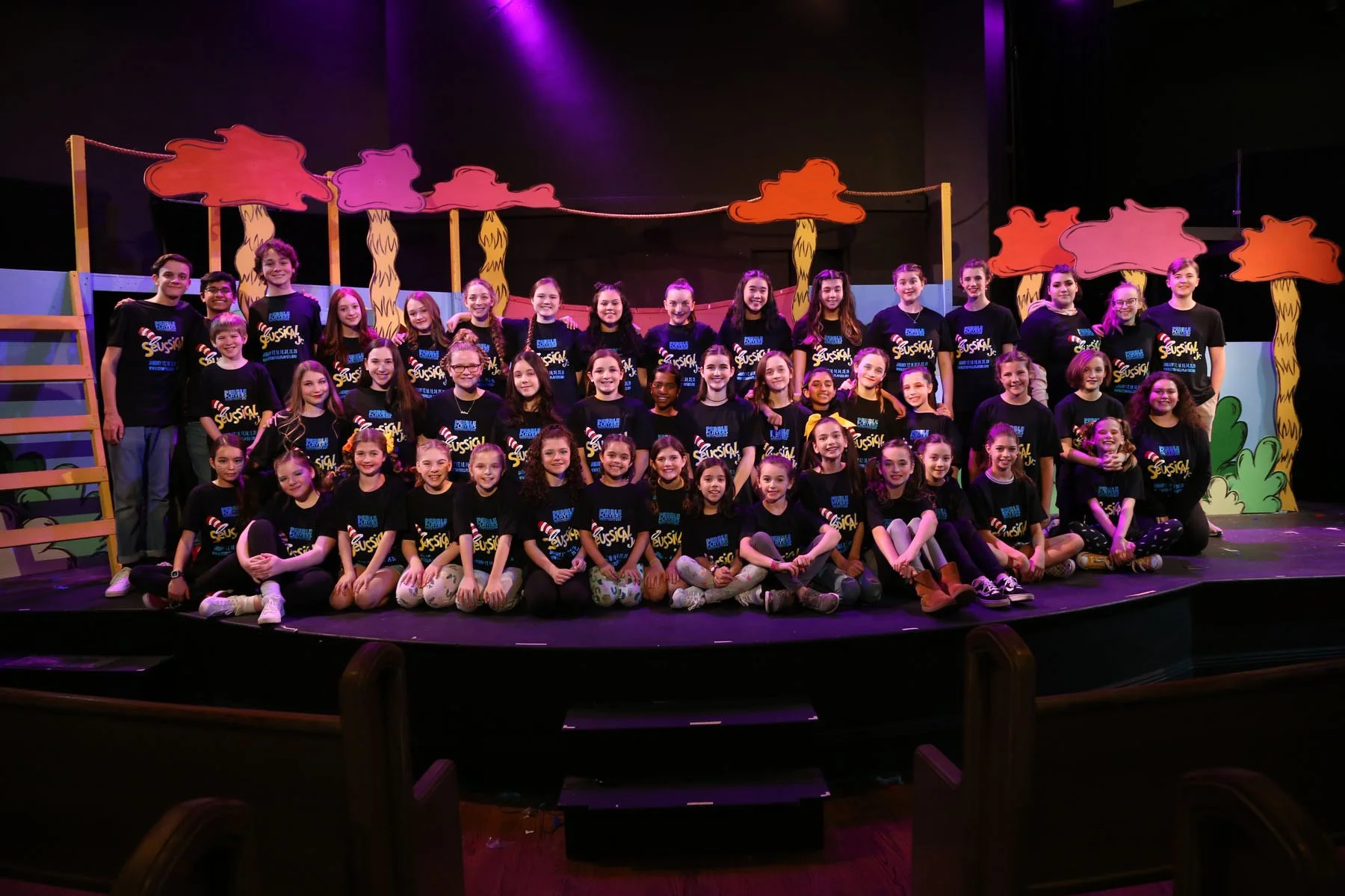 Group of children and teenagers on stage during a theatrical performance of Seussical, with colorful set pieces of trees in the background.