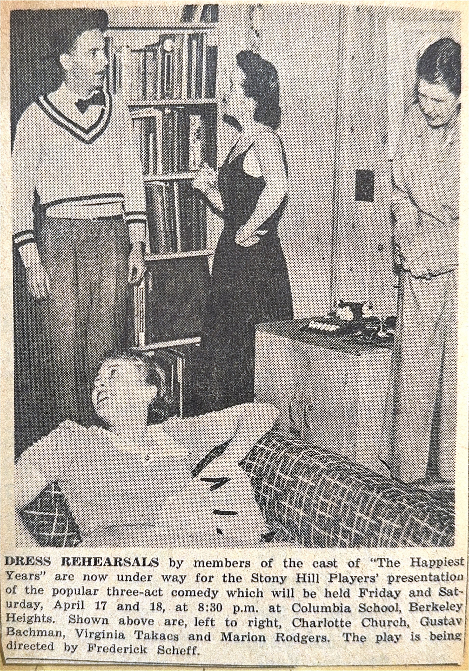 Black and white photo showing four people in a room. Two women are standing and three men, one shirtless, are sitting. The woman on the left is dressed in a sweater and bow tie. The woman in the middle is wearing a sleeveless dress. The woman on the 