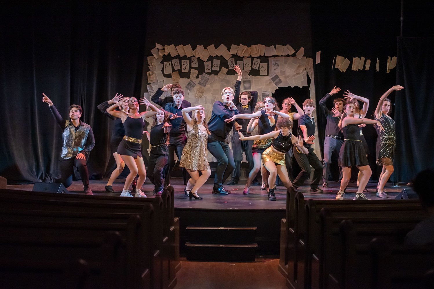 A group of young performers dancing on a stage with a backdrop of books and papers. They are dressed in a mix of casual and shiny costumes, striking energetic poses. Performance of Page to Stage cabaret.