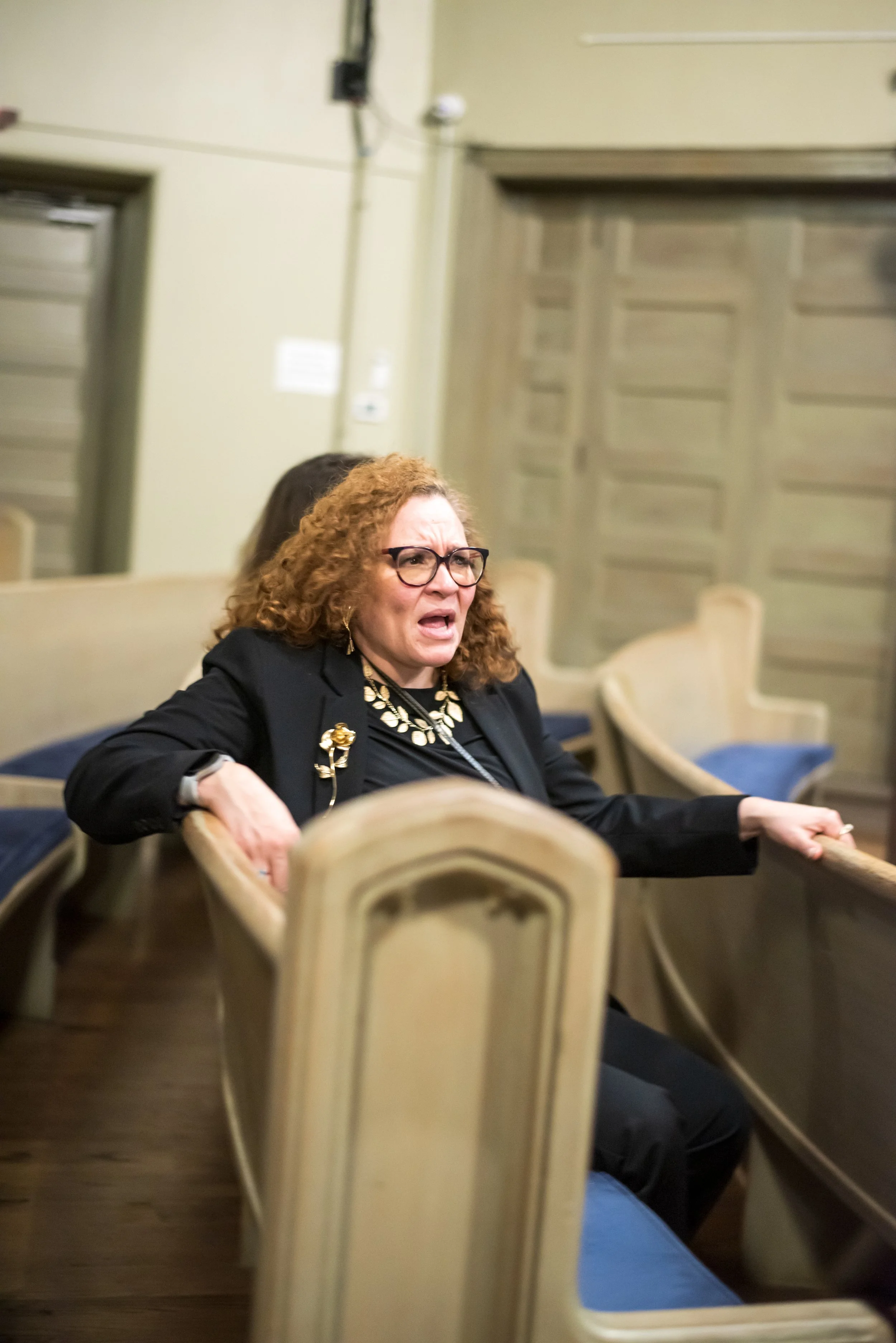 A woman with curly red hair, wearing glasses, a black blazer, and a large necklace, is sitting in a church pew with an emotional expression, possibly crying or upset.