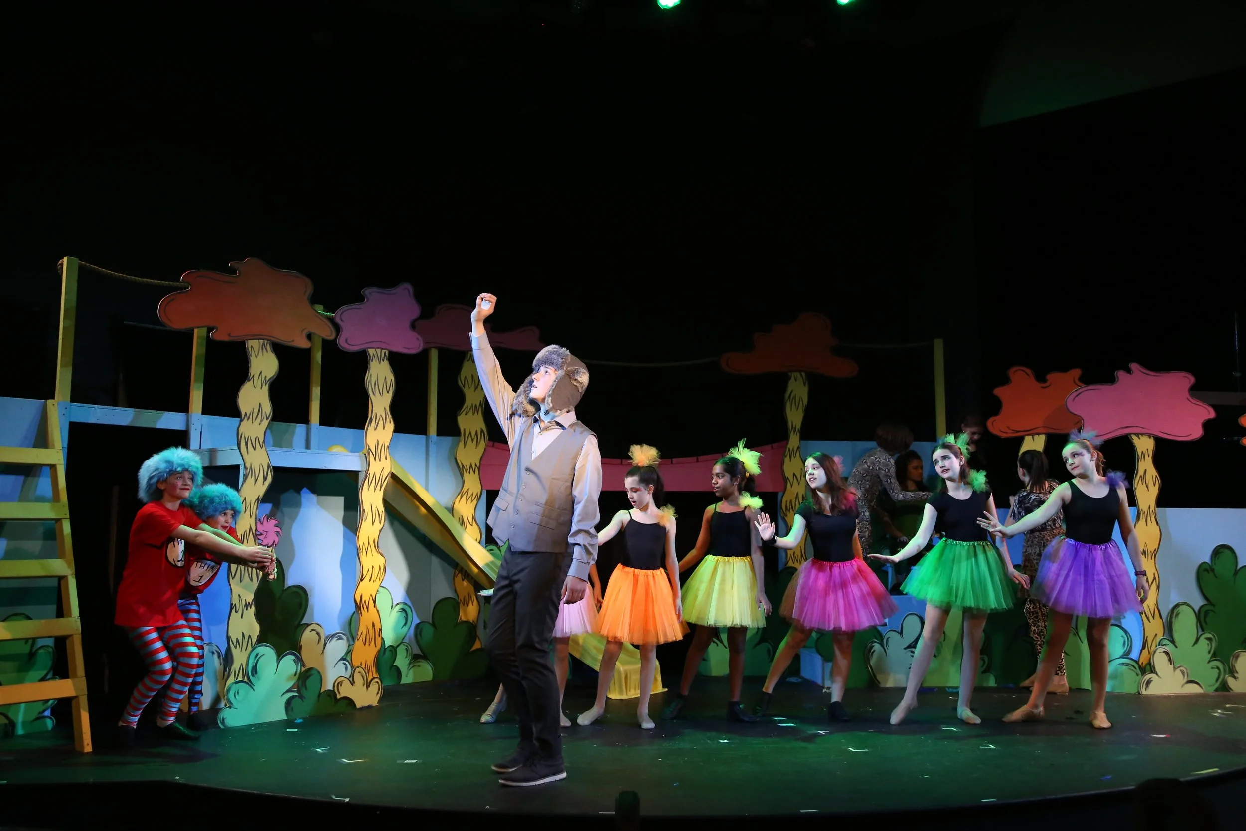 Children performing in Seussical Jr with a backdrop of cartoon-like trees and bushes, dressed in bright costumes including tutus and animal-themed accessories.