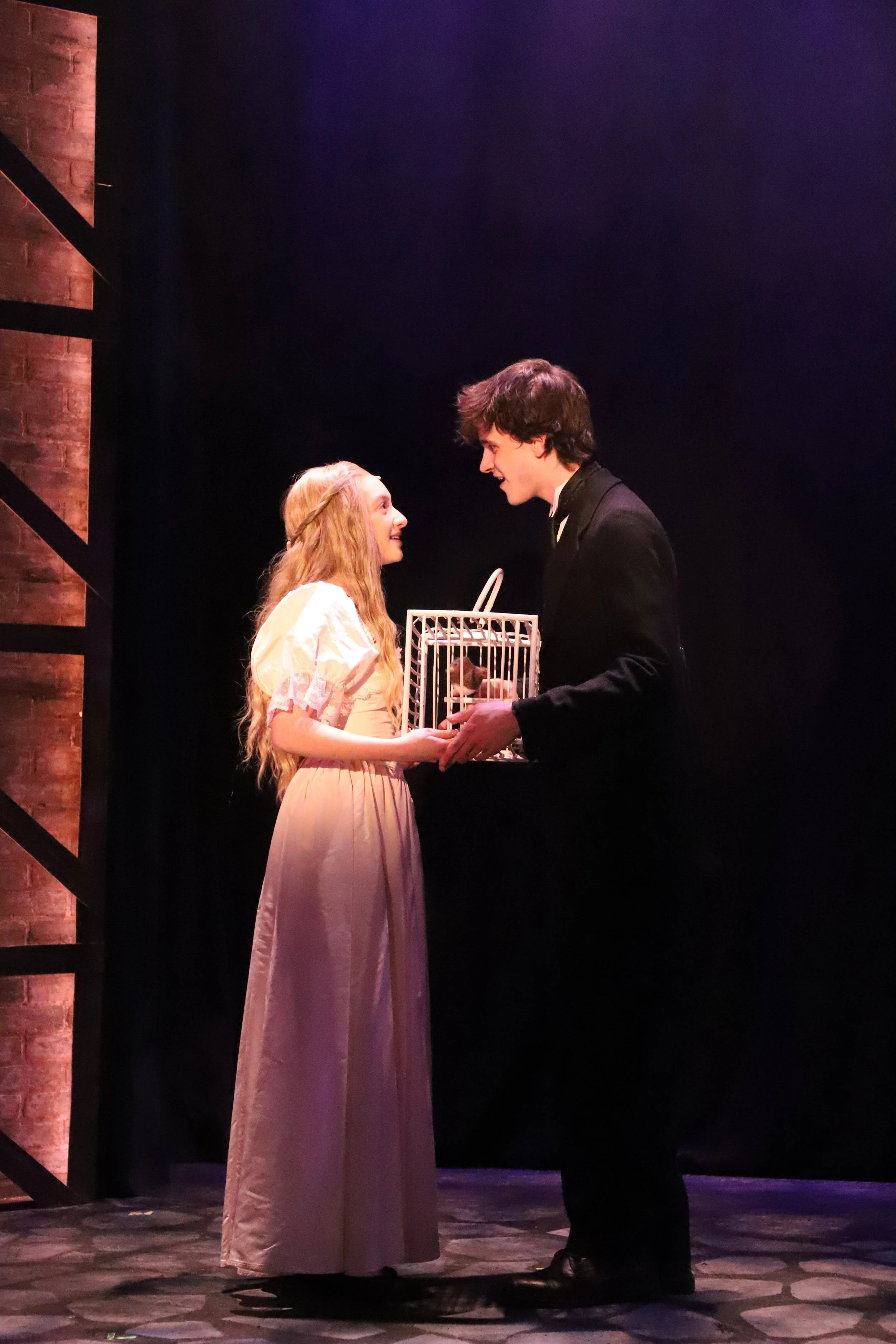 Two actors performing on stage, a woman with long, wavy blonde hair in a light-colored dress, and a man with dark, curly hair in a dark suit, are holding a birdcage together, engaging in a theatrical scene.
