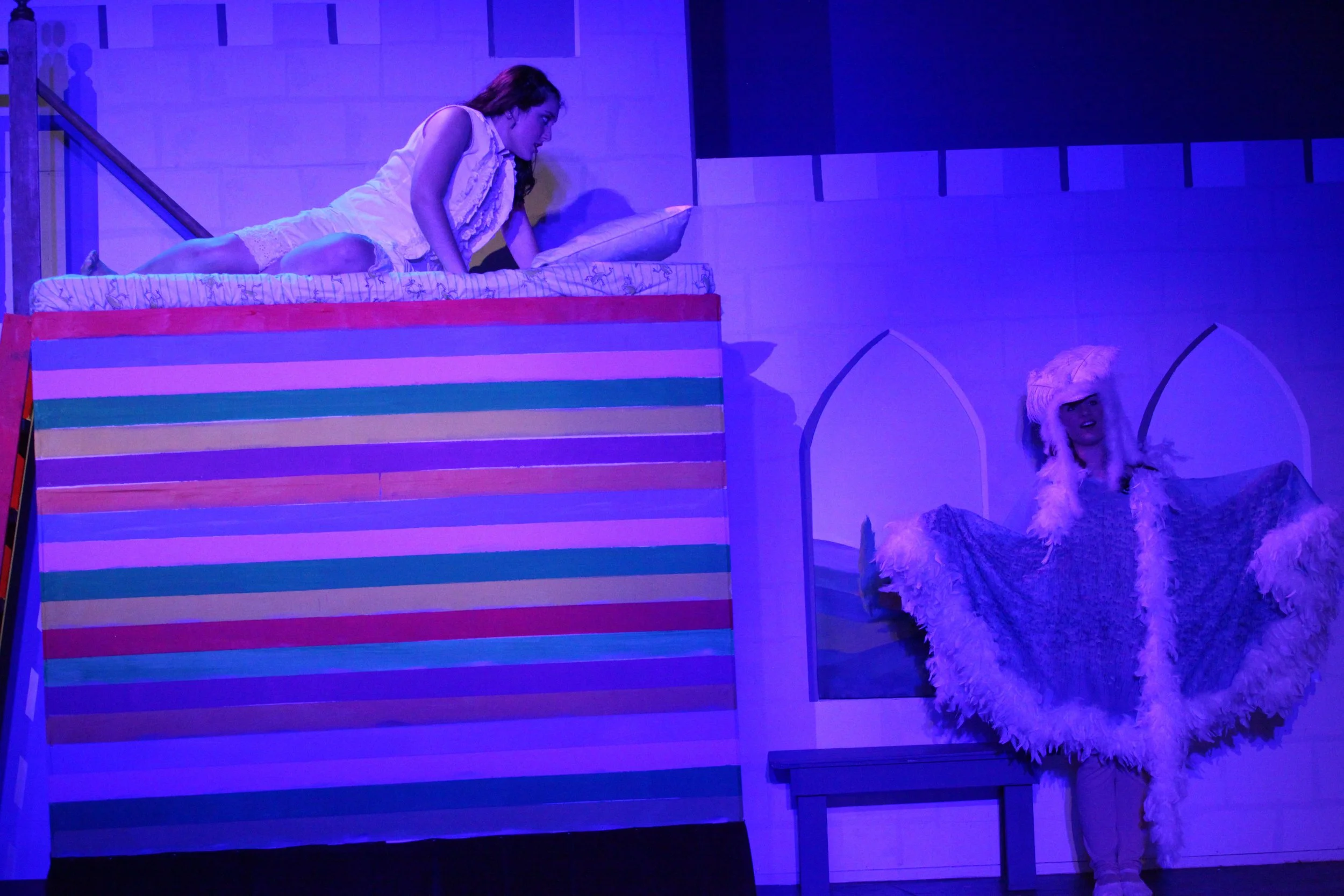 A theatrical stage scene featuring a woman lying on a bed, dressed in white, looking towards another woman who is standing and dressed in a fluffy costume, inside a stylized room set with colorful stripes and painted arches.