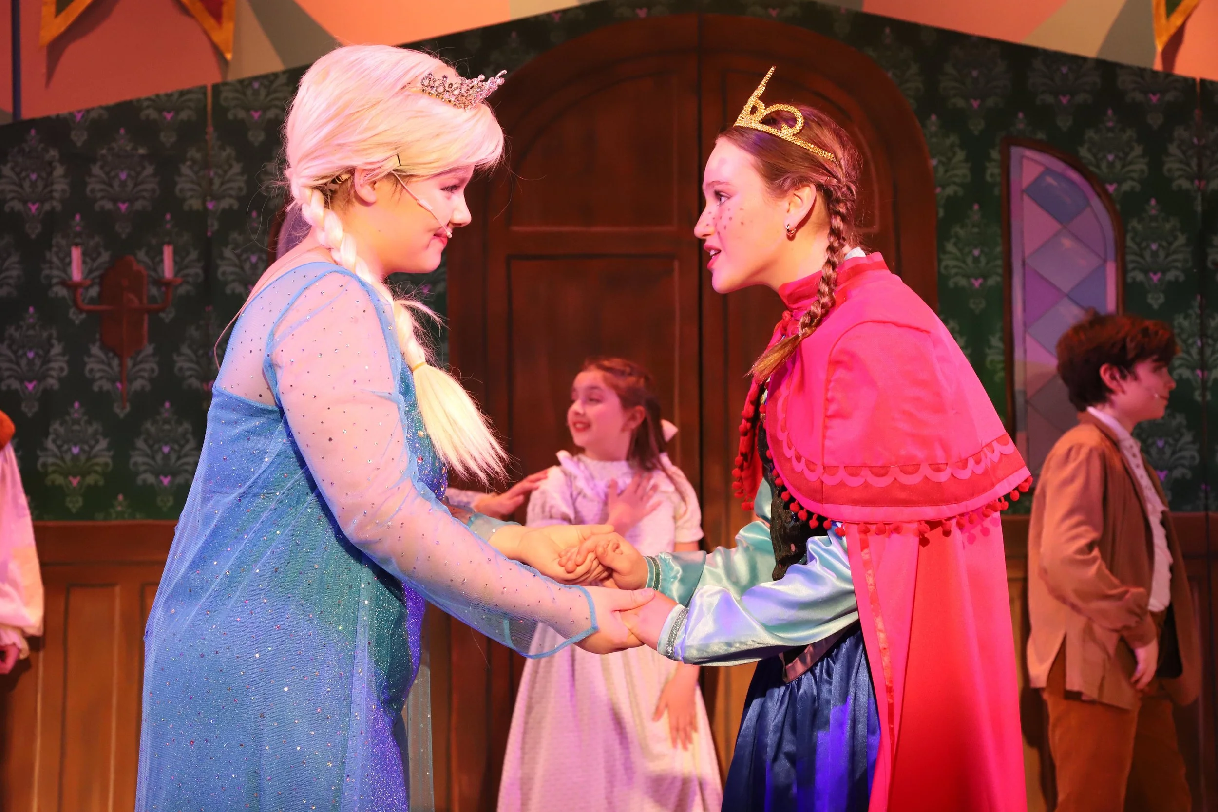 Two young girls dressed as Anna and Elsa from Frozen Jr. holding hands and smiling at each other on stage during a play, with other children in costume in the background.