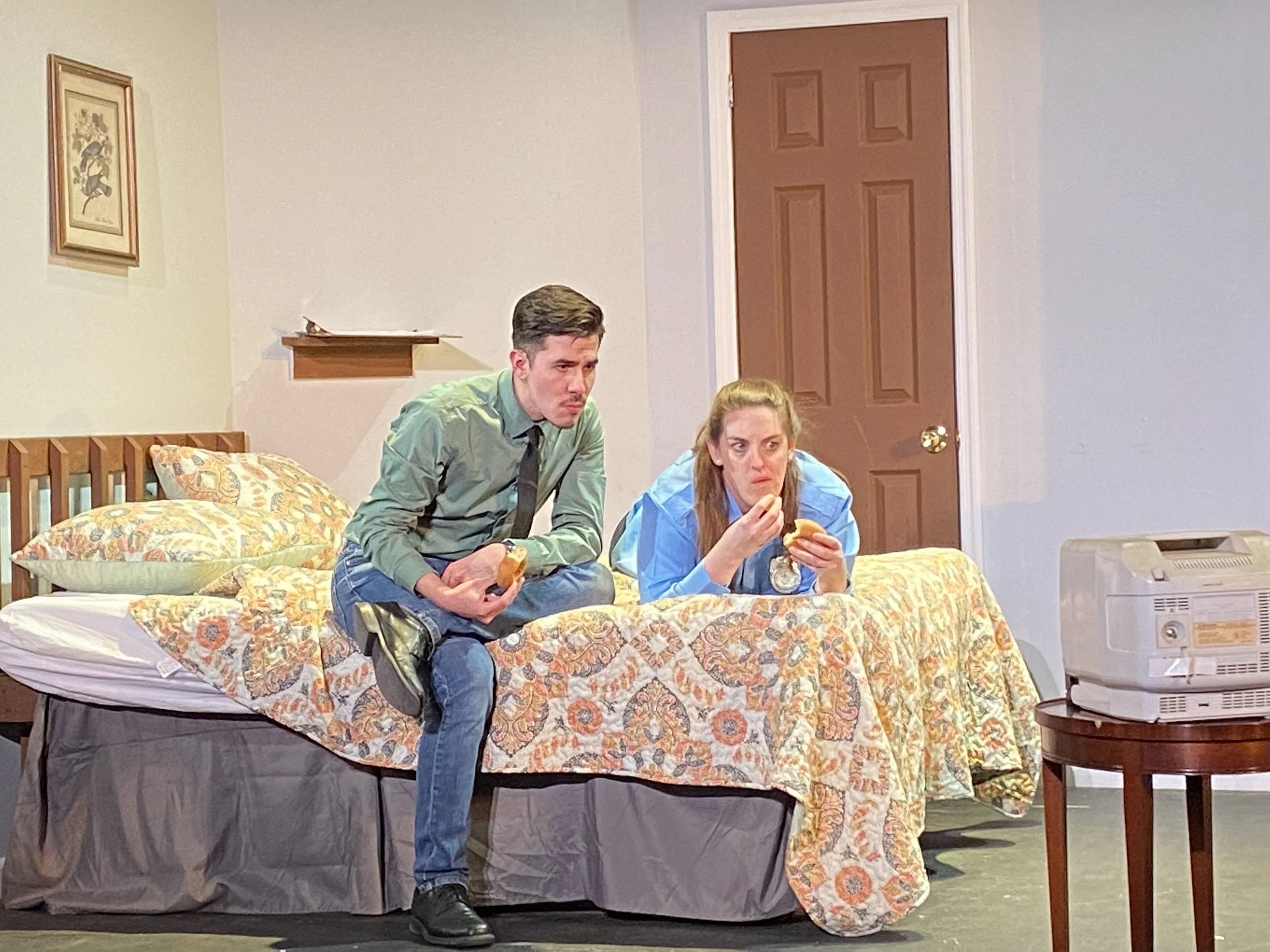 A man and woman sitting on a bed with a floral bedspread, eating fast food in a room with a brown door, a framed picture on the wall, and an old television on a wooden table.