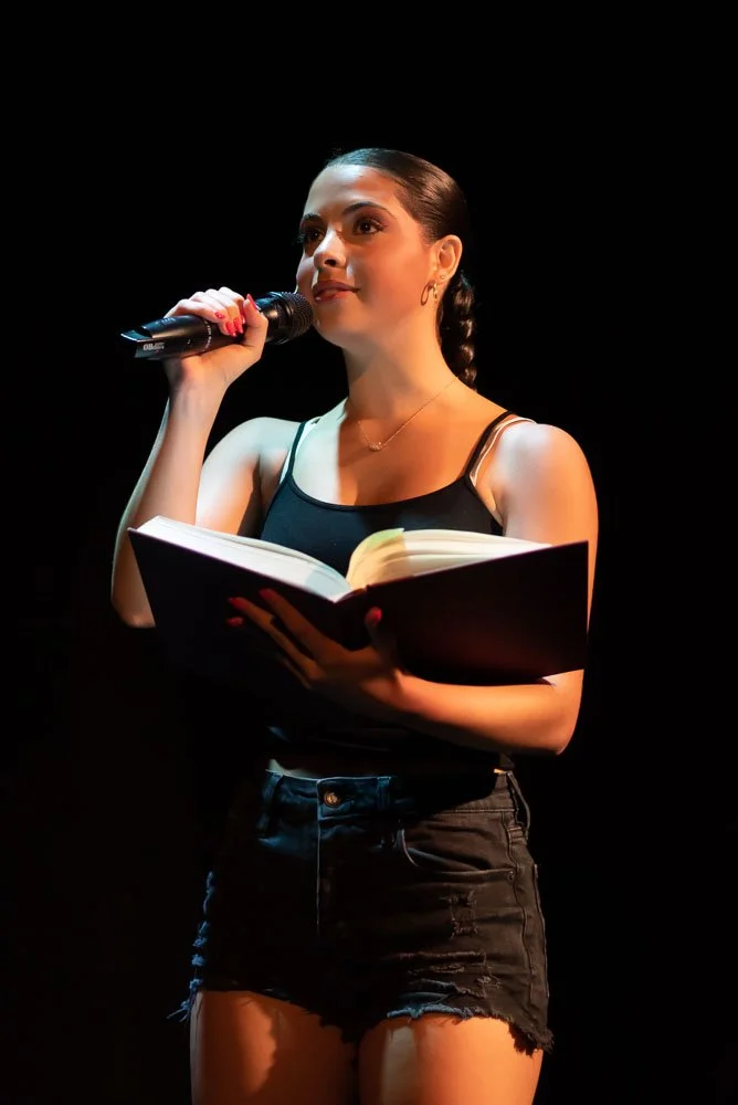 Young woman on stage holding a book and speaking into a microphone, wearing a black tank top and black denim shorts, with a dark background. Performance of Page to Stage cabaret.