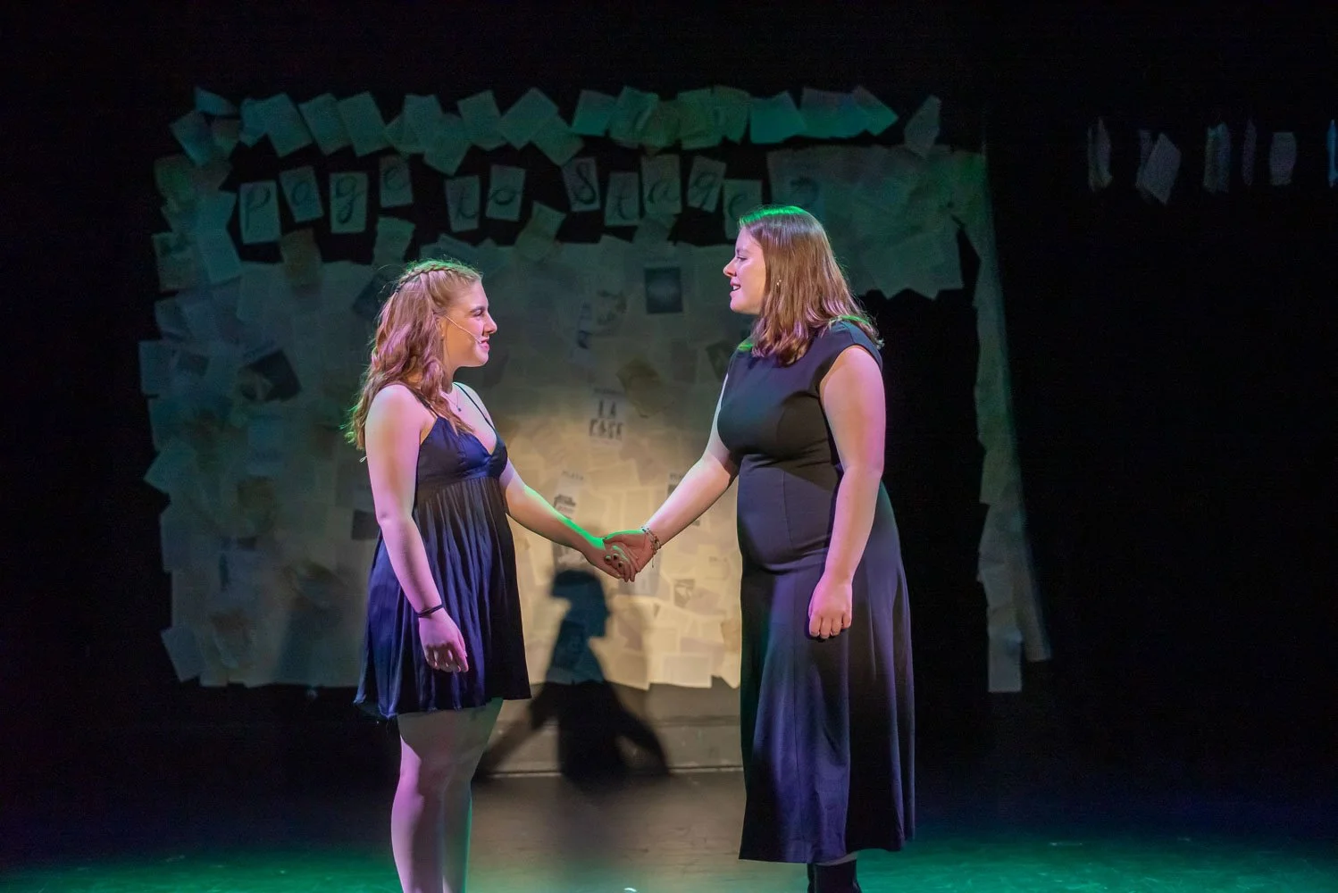 Two women on stage, holding hands and smiling at each other during a theatrical performance of Page to Stage cabaret., with a backdrop of paper and sign decorations.